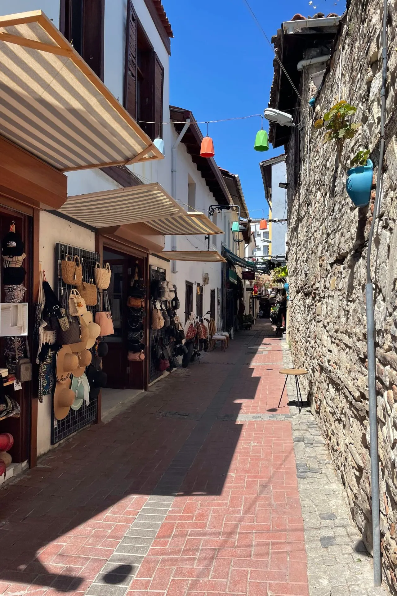 Smalle winkelstraat in Kuşadası met kleurrijke hoeden, tassen en schaduwrijke luifels aan de gevels.