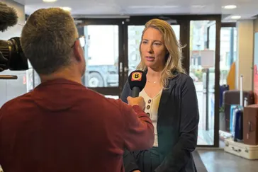 Een journalist van Omroep Brabant interviewt Michelle de Wit, eigenaresse van Travelcenter de Wit, bij de ingang van het reisbureau. De journalist houdt een microfoon met het logo van Omroep Brabant vast, terwijl een cameraman het interview filmt. Op de achtergrond zijn koffers en de glazen entree van het kantoor zichtbaar.