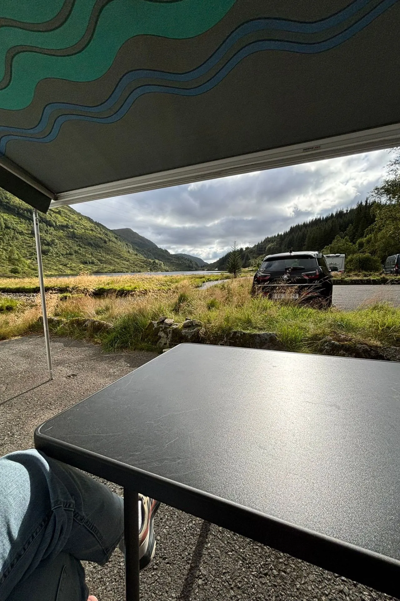 Uitzicht vanaf een campingplek onder een luifel, met een zwarte tafel op de voorgrond. Op de achtergrond een geparkeerde auto, gras en een rivier, omringd door groene bergen en bossen onder een deels bewolkte lucht.