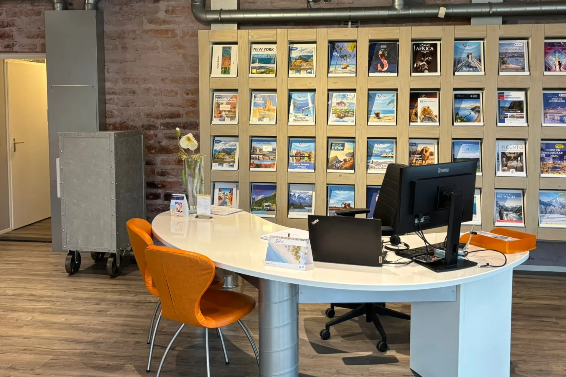 Binnenkant van het filiaal in Asten met een wit afgerond bureau, oranje stoelen en een wand vol reisbrochures van onder andere TUI.
