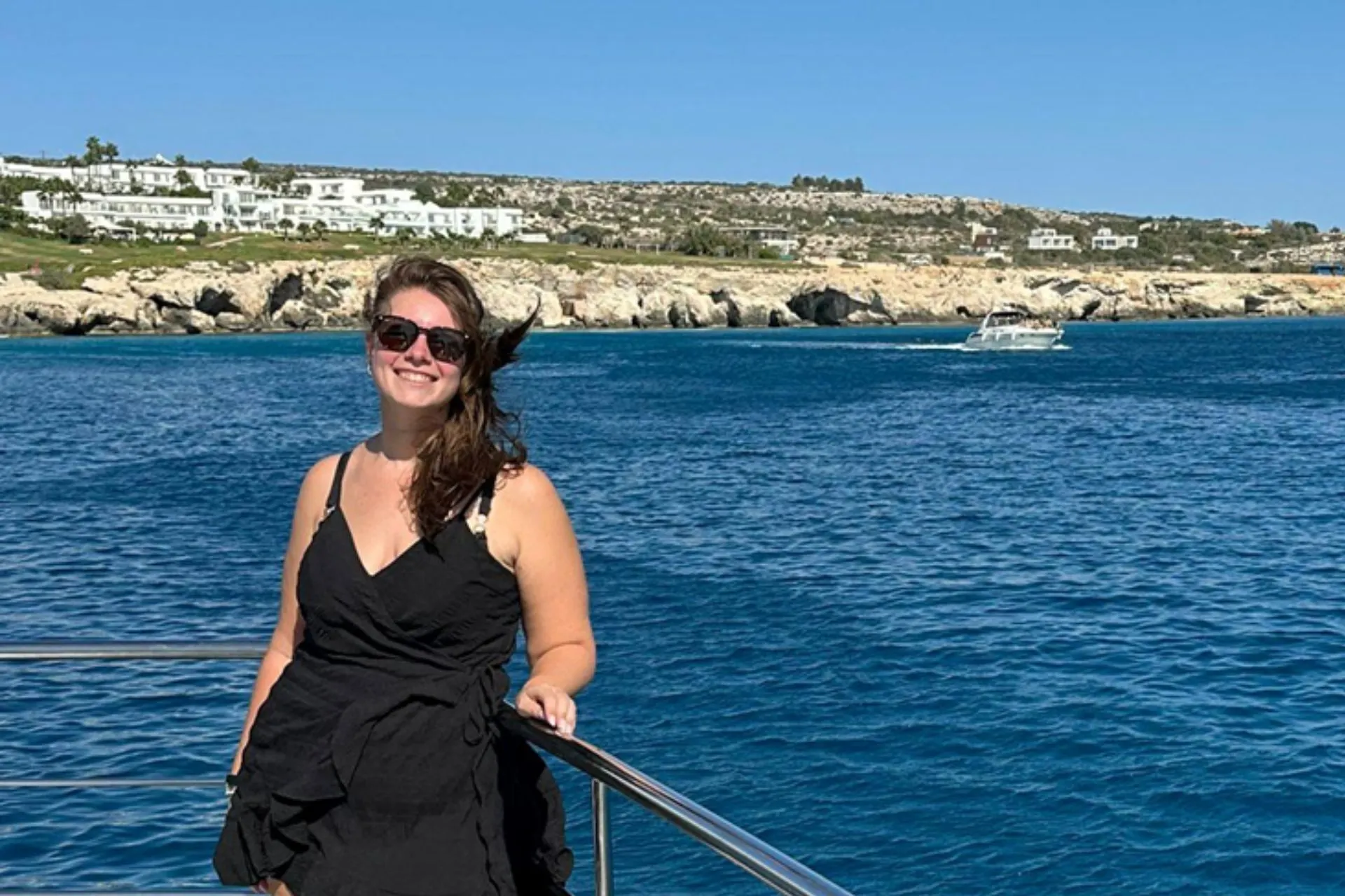 Persoon staat op het dek van een boot bij een railing, met uitzicht op helderblauw water en een kustlijn met witte gebouwen in de verte.