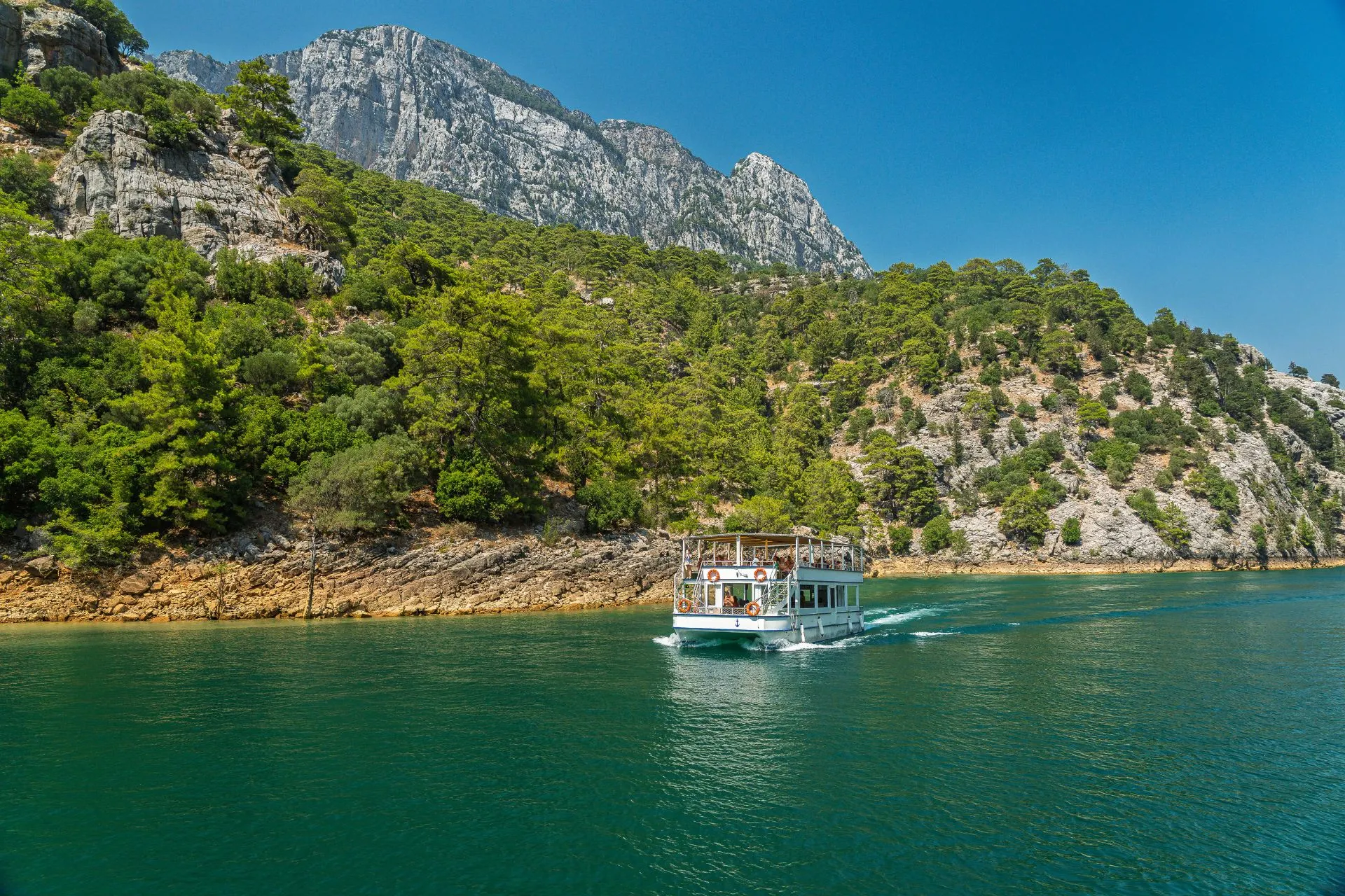 Rondvaartboot op het helderblauwe water van de Green Canyon nabij Manavgat en Side. Omringd door groene heuvels, dennenbossen en indrukwekkende bergtoppen biedt deze excursie een unieke manier om de Turkse natuur te ontdekken. Een van de populairste uitstapjes in de omgeving van Side.