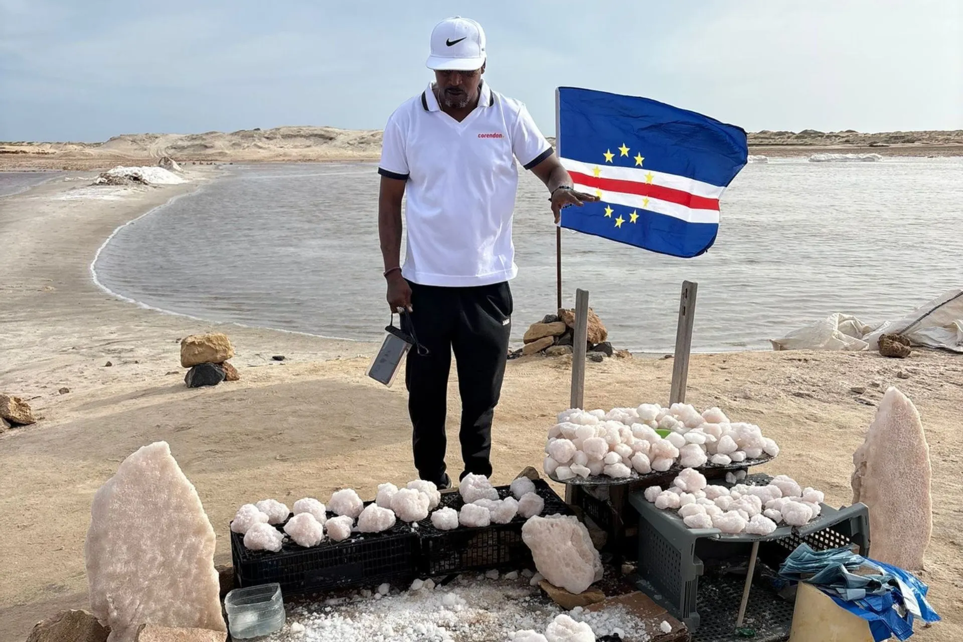 Een persoon staat bij bakken vol grote zoutkristallen naast de zoutvlaktes, terwijl een Kaapverdische vlag aan een stok naast hem wappert.