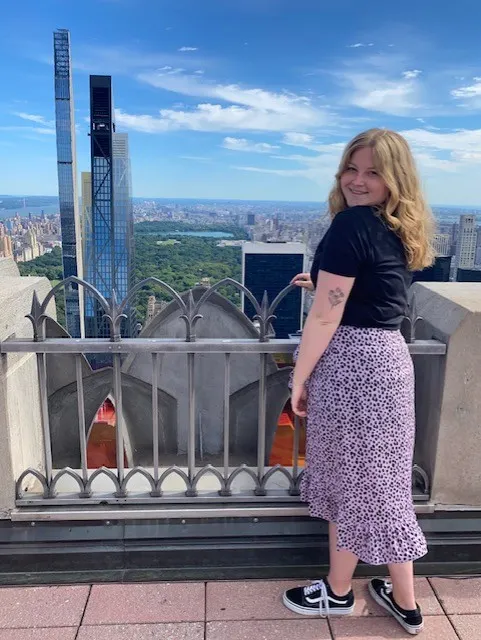 Een blonde vrouw kijkt uit over de stad, met een panoramisch uitzicht op de skyline van New York en de iconische wolkenkrabbers op de achtergrond.