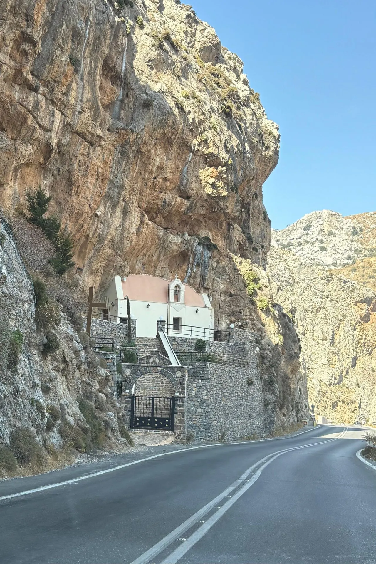 Kleine witte kapel met terracotta dak ingebouwd in een rotswand langs een bergweg op Kreta, omgeven door steile kliffen.