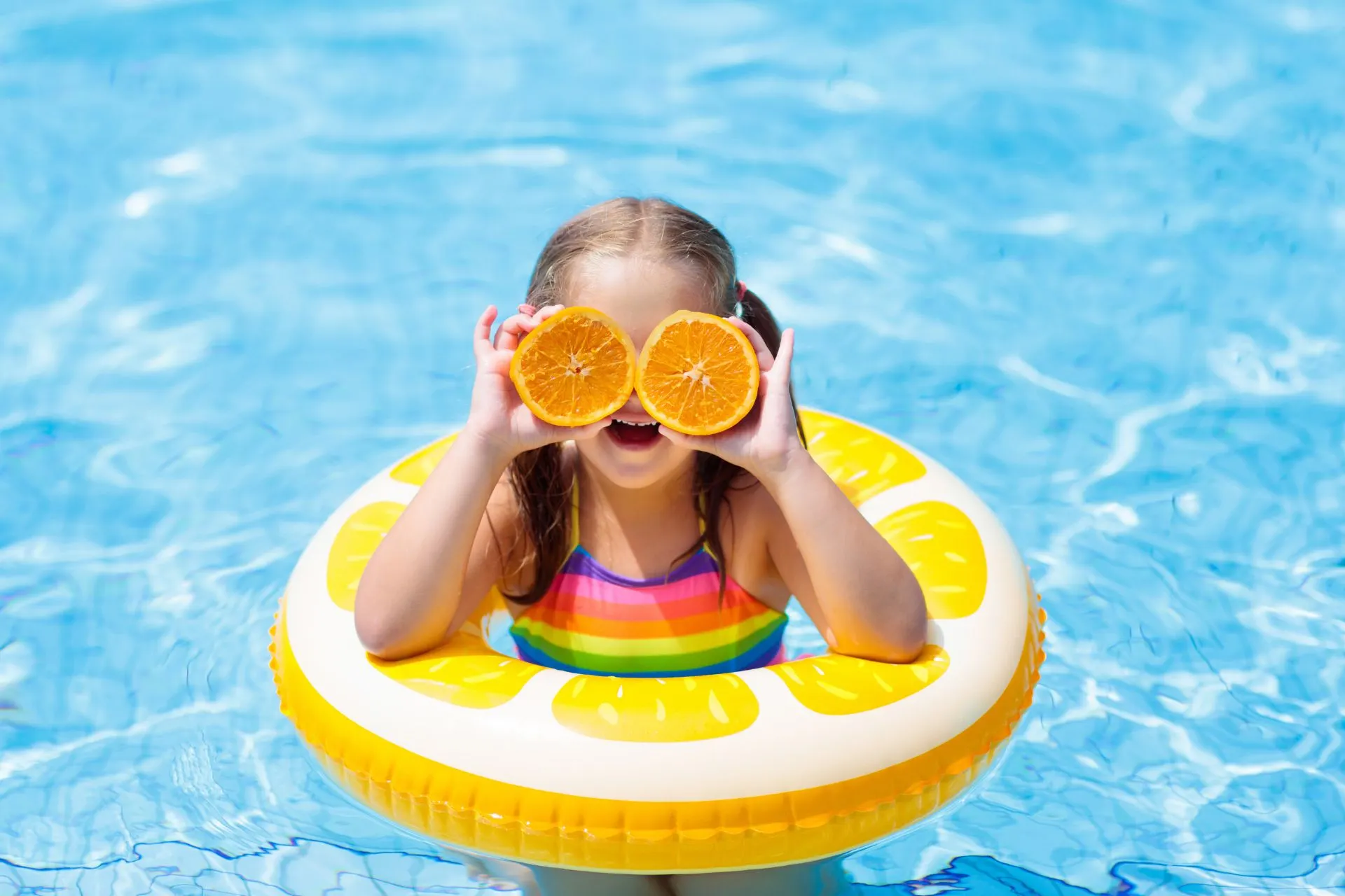 Kind speelt vrolijk in een helderblauw zwembad met een kleurrijke opblaasband in citrusdesign en houdt twee halve sinaasappels vast. Deze kindvriendelijke all-inclusive vakantie combineert plezier, zon en verfrissing. Perfect voor gezinnen die op zoek zijn naar een zorgeloze vakantie vol waterpret en ontspanning in een veilige en comfortabele omgeving.