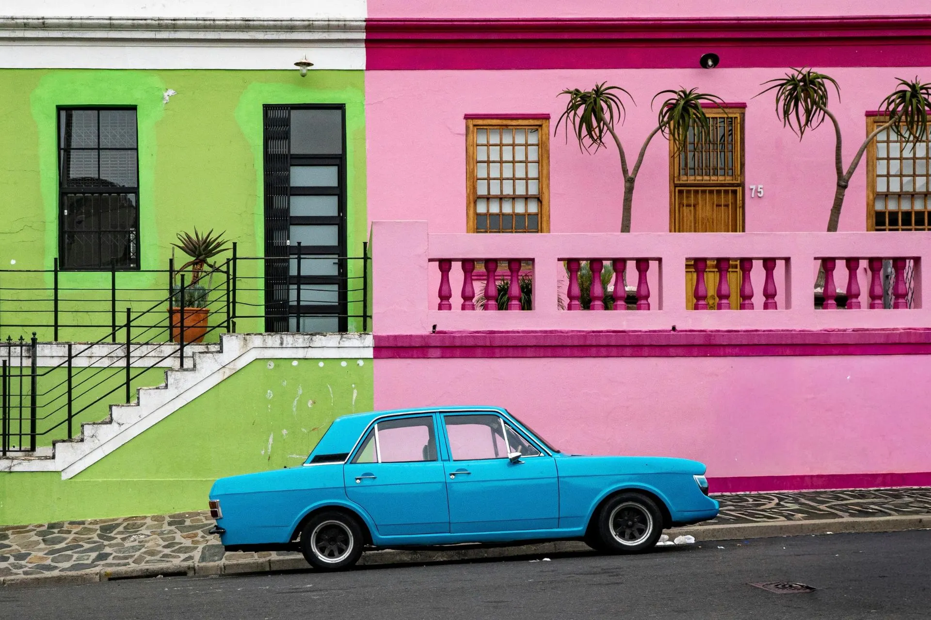 Levendige straat met felgekleurde gevels in groen en roze, gescheiden door een verticale lijn, en een opvallende blauwe klassieke auto geparkeerd op de voorgrond. De architectuur is eenvoudig maar charmant, met trappen, metalen hekwerken en tropische planten die een exotische sfeer creëren. Dit beeld straalt authenticiteit en kleur uit, typerend voor bestemmingen zoals Kaapstad, een van de beste winterzon bestemmingen voor reizigers die op zoek zijn naar zon, cultuur en unieke ervaringen. Terwijl in Europa de winter heerst, biedt deze plek een perfecte zonvakantie in december of een zonvakantie in januari, met aangename temperaturen en volop zon. Voor iedereen die zich afvraagt: waar is het warm in januari? Zuid-Afrika is het antwoord. Naast prachtige stranden en natuur, vind je hier ook kleurrijke wijken zoals Bo-Kaap, ideaal voor fotografie en cultuur. Ontdek waarom zoveel reizigers deze plek kiezen als een van de beste winterzon bestemmingen en plan jouw perfecte zonvakantie in december of januari op een locatie waar het altijd warm en gastvrij is.