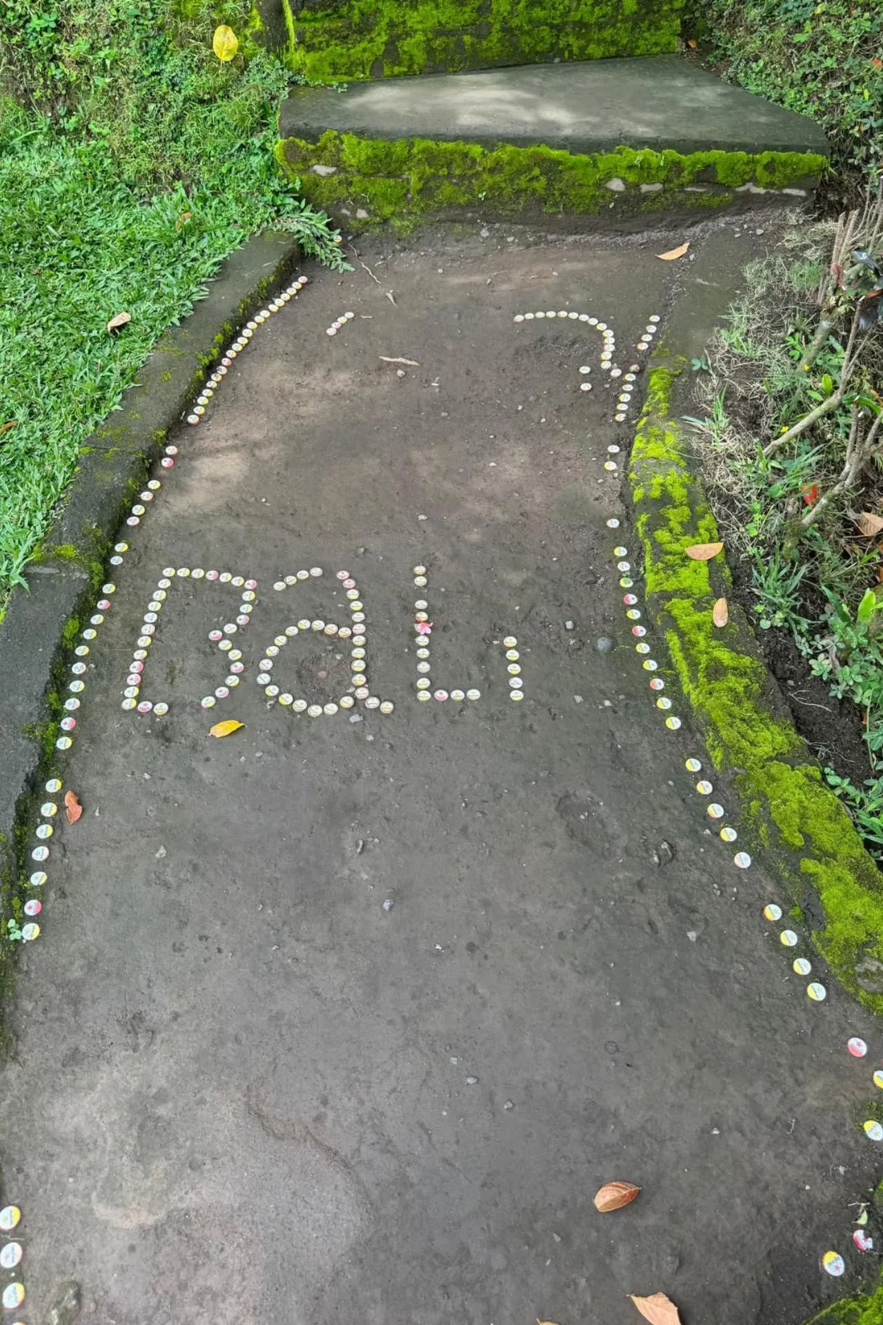 Pad op Bali met kleurrijke steentjes die het woord 'Bali' vormen, omlijst door mosrijke trappen