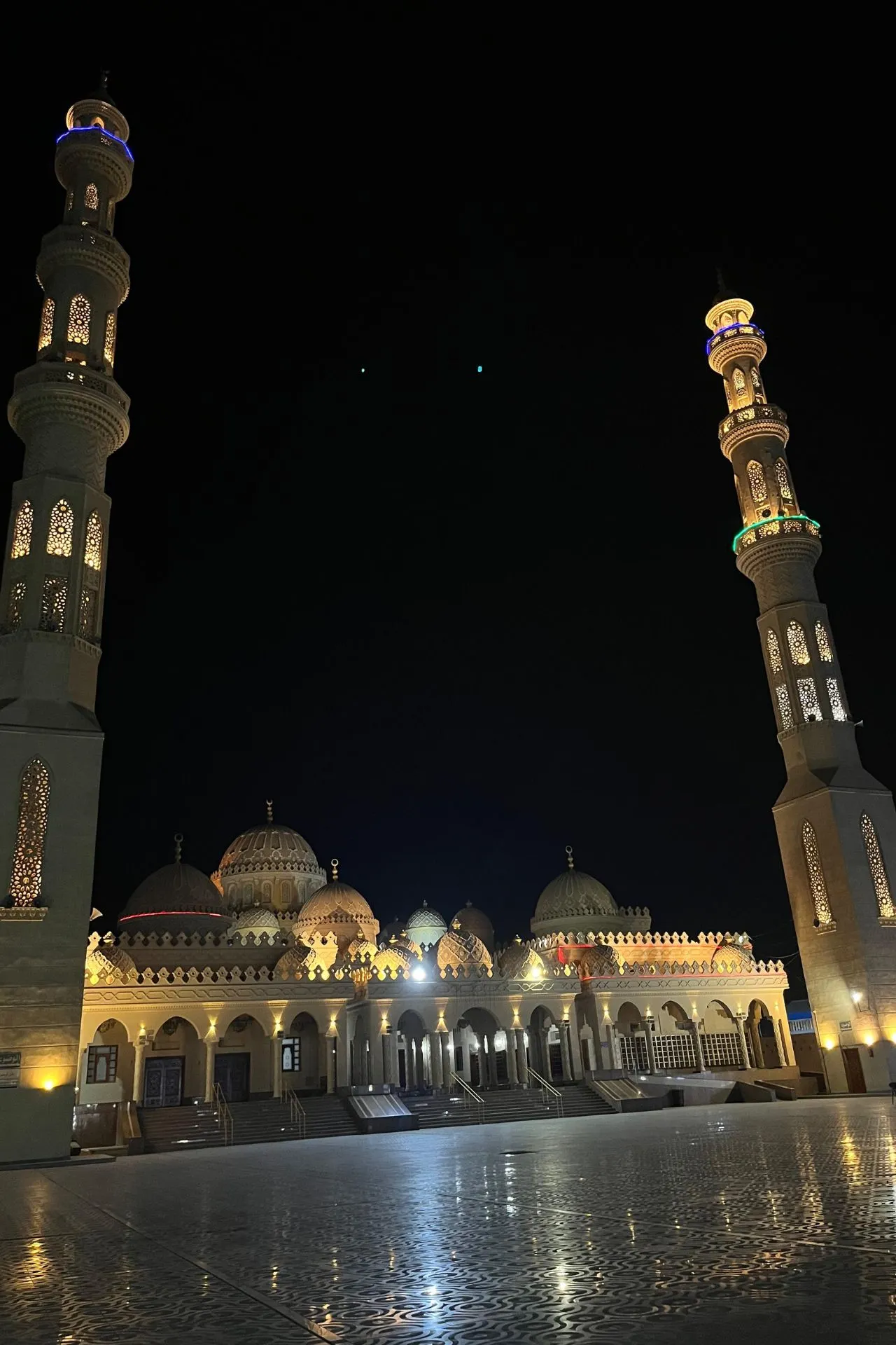 Indrukwekkend nachtbeeld van een grote moskee met prachtig verlichte minaretten en gedetailleerde architectuur. De moskee heeft meerdere koepels en sierlijke bogen, terwijl de minaretten rijkelijk versierd zijn met patronen en lichtaccenten die opvallen tegen de donkere nachtelijke hemel. De voorgrond toont een glanzend betegeld plein dat de verlichting weerspiegelt, wat een serene en majestueuze sfeer creëert. Ideaal voor promotie van culturele bezienswaardigheden, religieuze architectuur en nachtelijke stadsfotografie in Egypte.