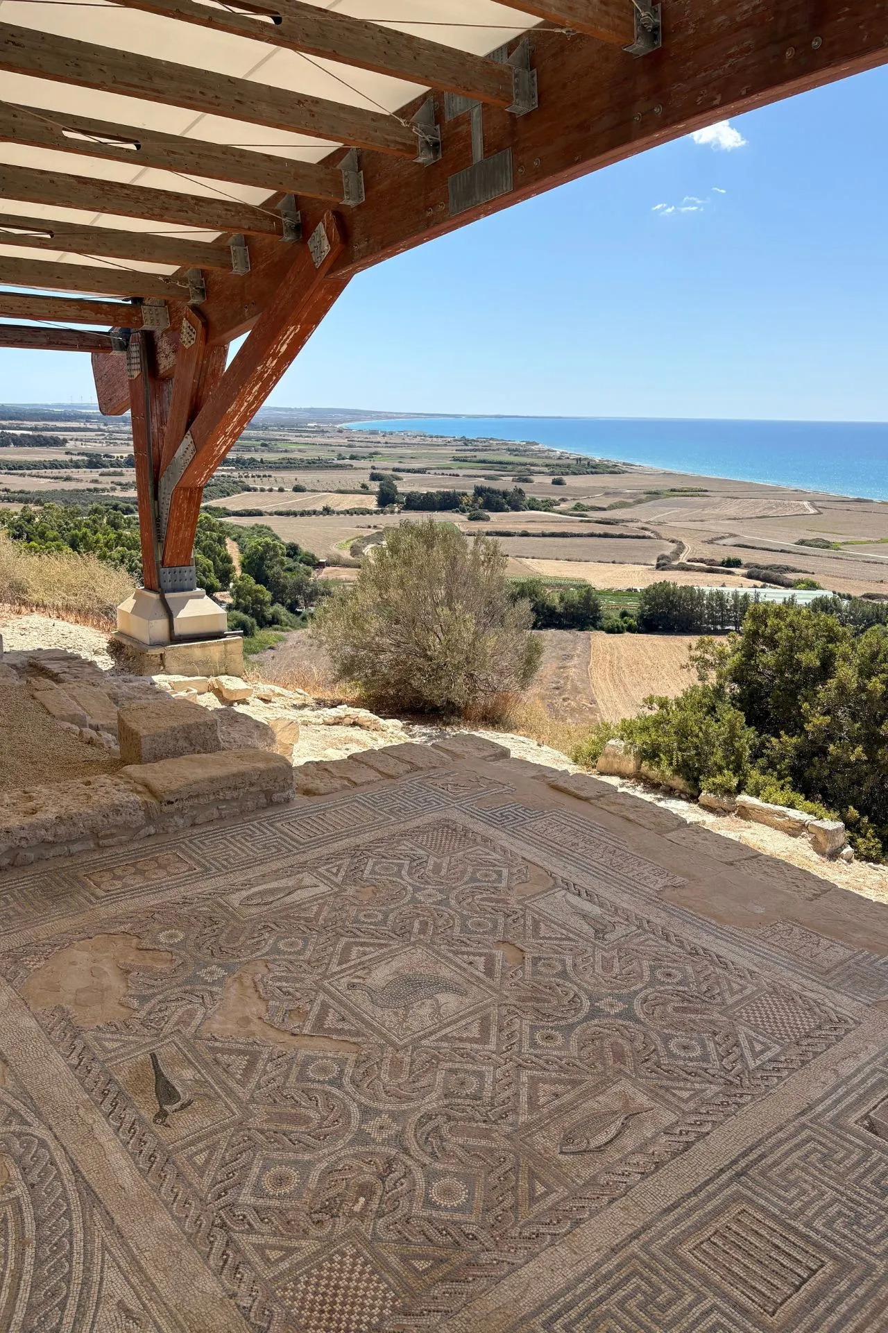 Oude mozaïekvloer onder een houten overkapping bij de archeologische site van Kourion, met uitzicht op landbouwvelden en de Middellandse Zee.