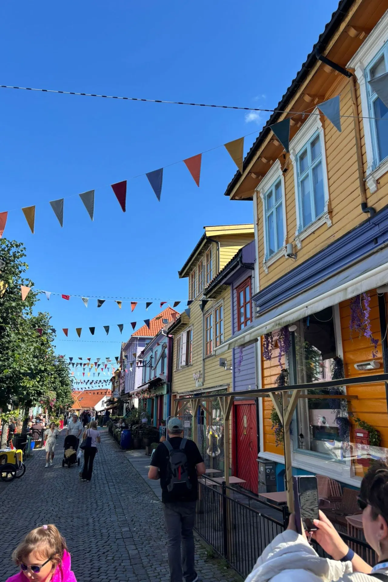 Gezellige straat met kleurrijke houten huizen en vlaggetjes boven de weg, onder een heldere blauwe lucht. Mensen wandelen langs winkels en cafés in een charmante, historische omgeving.