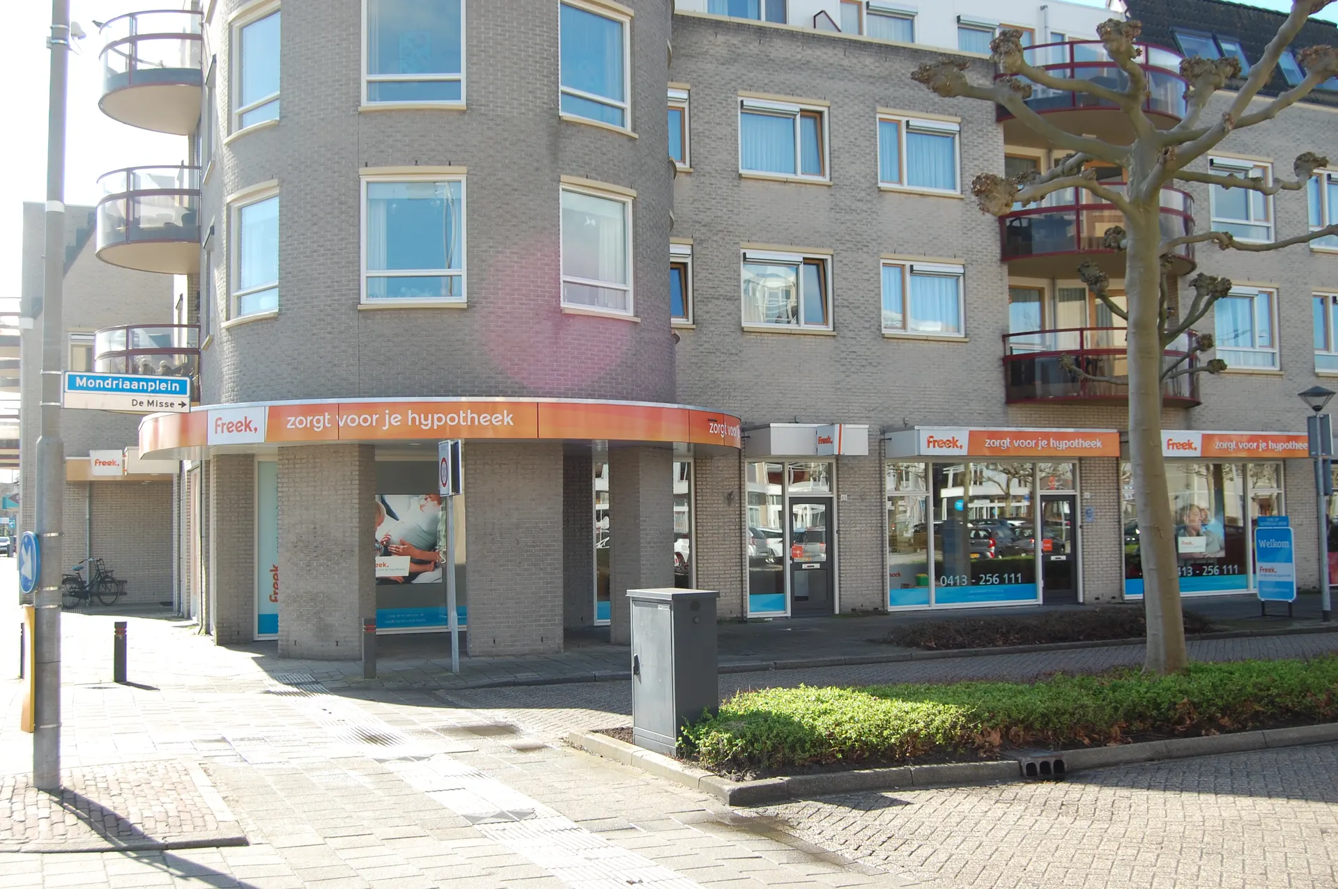  "Close-up van een modern hoekpand met lichte bakstenen gevel, grote ramen en oranje gevelstroken met de tekst ‘zorgt voor je hypotheek’. Op de begane grond bevindt zich een commerciële ruimte, ideaal voor financiële dienstverlening, kantoren of retail. Het gebouw heeft een eigentijdse architectuur met ronde balkons en een stedelijke uitstraling. Perfecte locatie voor zakelijke activiteiten, goed bereikbaar en gelegen in een levendige wijk met voetgangerszone en parkeermogelijkheden."