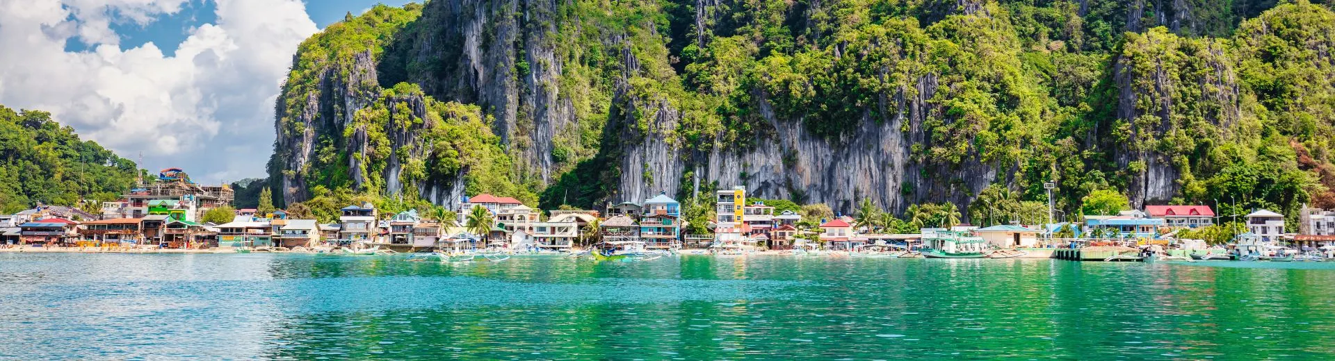 Uitzicht op een kustplaats in de Filipijnen met kleurrijke huizen langs het turquoise water, omringd door imposante kalksteenrotsen en weelderige groene vegetatie onder een deels bewolkte hemel.