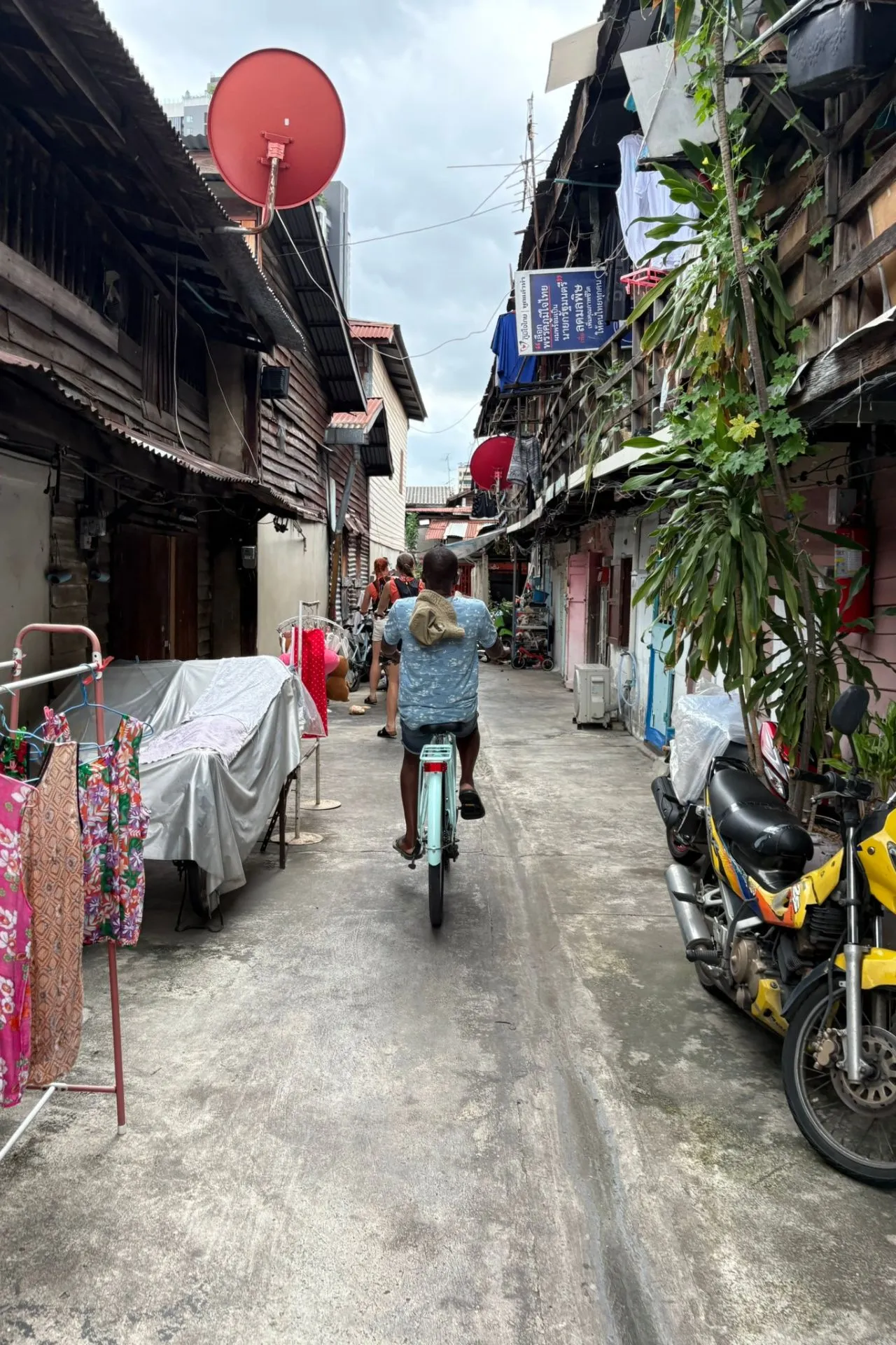 Achteraanzicht van een fietser die door een smalle straat rijdt, geflankeerd door houten huizen, kleurrijke waslijnen en motorfietsen.