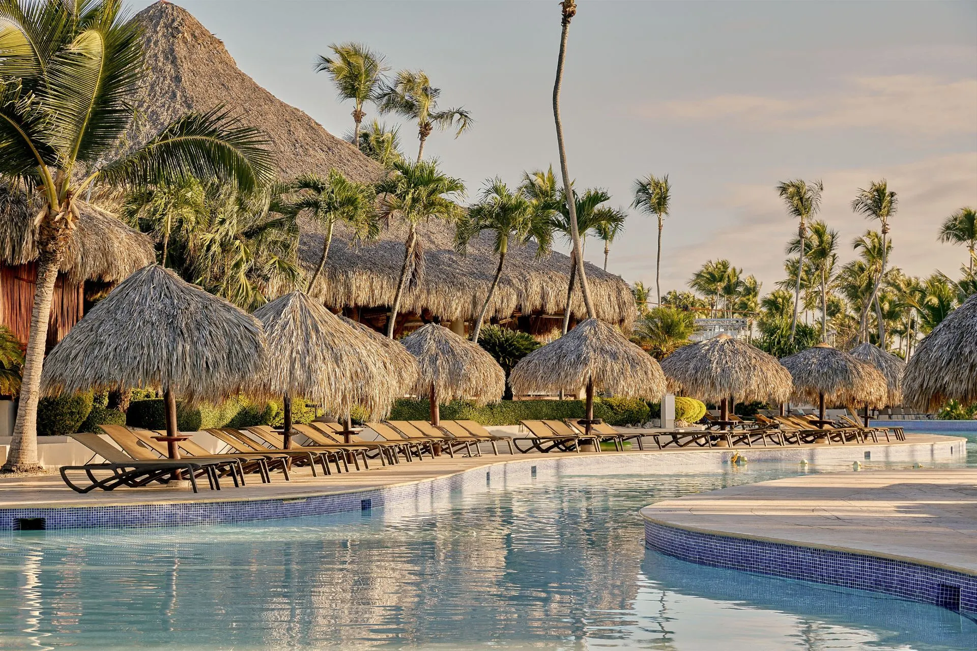 Zwembad bij Club Med Punta Cana omringd door rieten parasols en palmbomen. Ontspan tijdens jouw all-inclusive vakantie in de Caraïben met onbeperkte drankjes, wellness en sportactiviteiten.