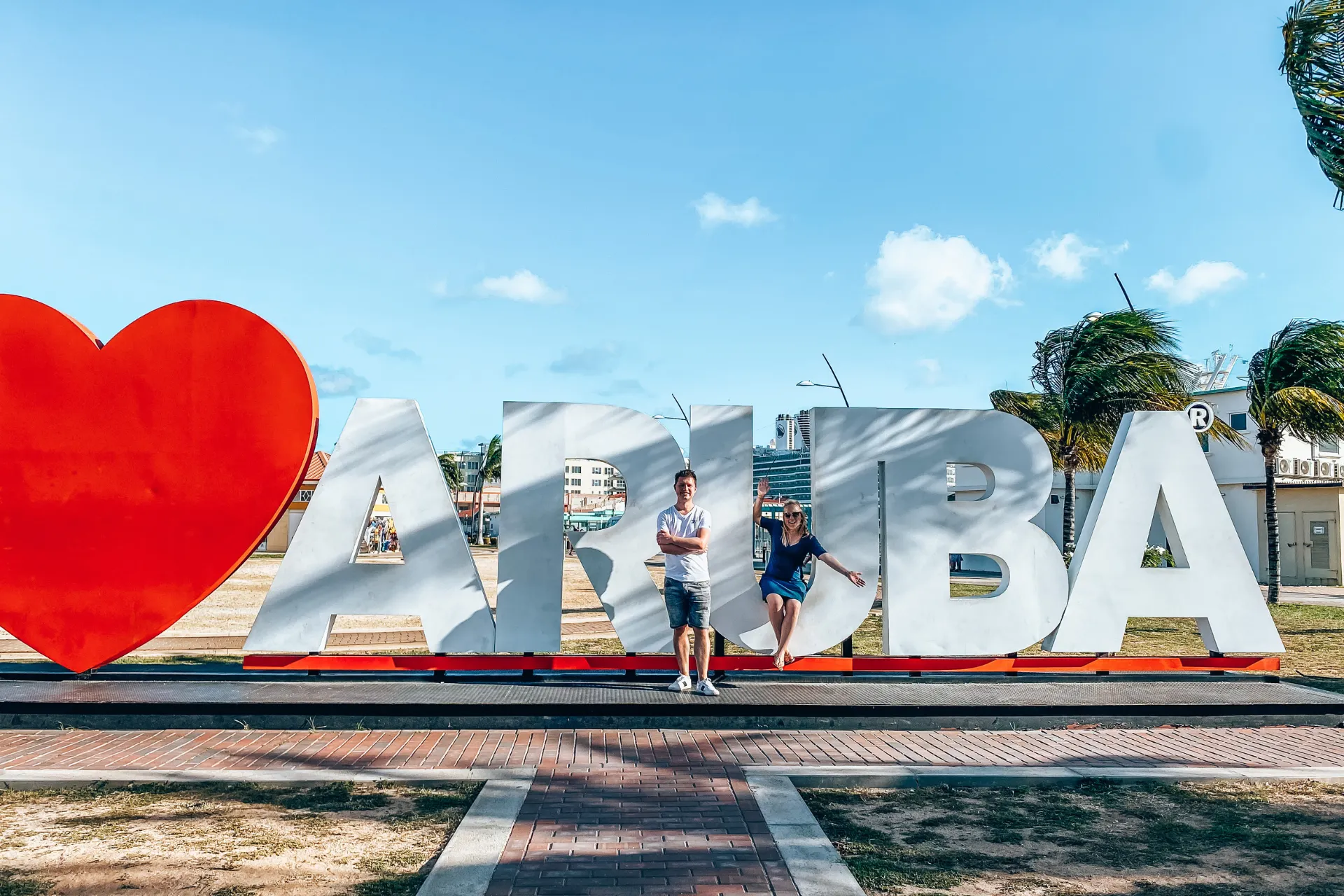 Grote witte letters met rode hartvorm die samen ‘I ❤️ Aruba’ vormen, geplaatst op een plein met betegelde ondergrond, palmbomen en blauwe lucht op de achtergrond, een iconische fotospot en populaire bezienswaardigheid op het eiland Aruba.