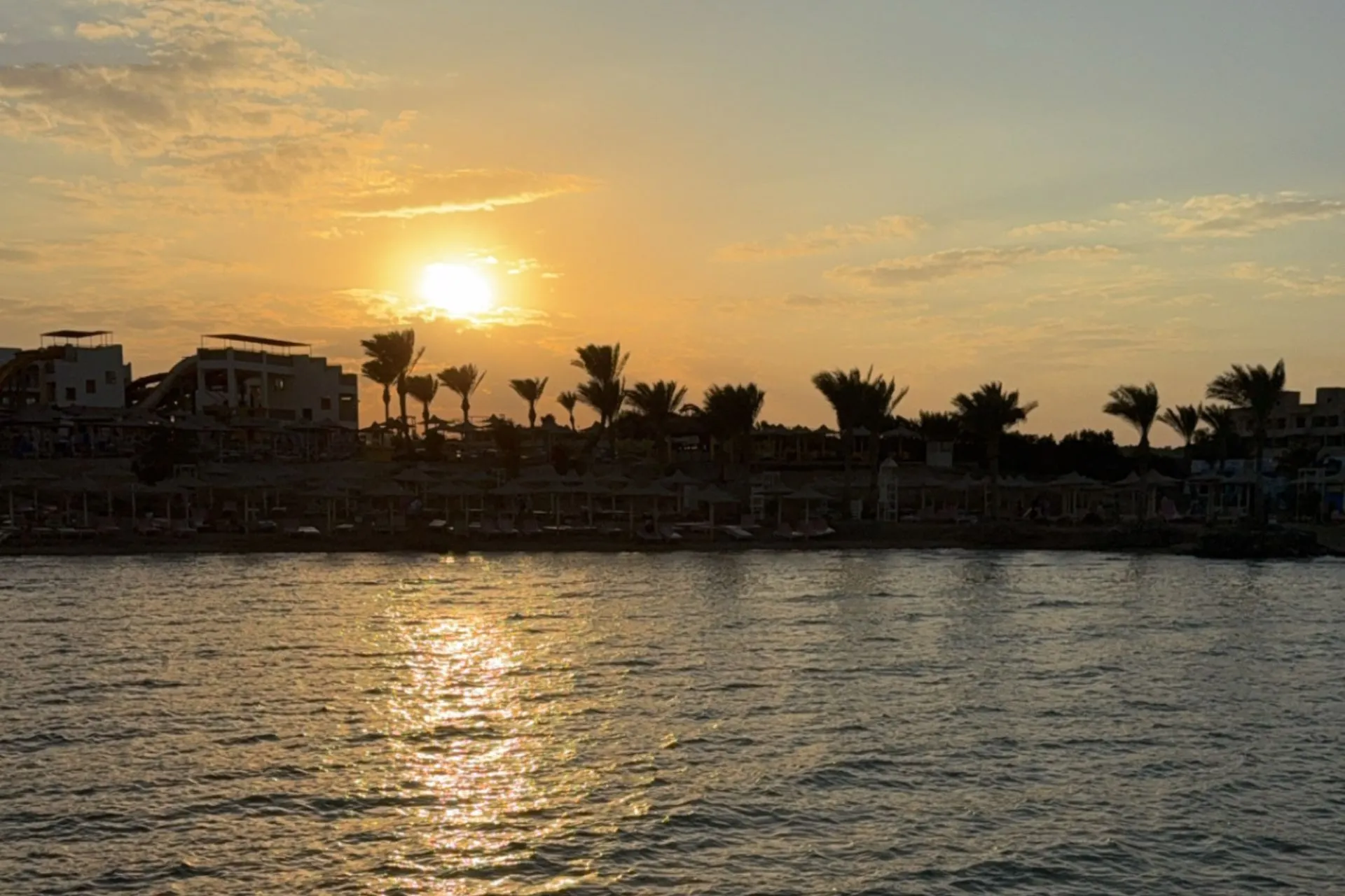 Zonsondergang boven een kustlijn met palmbomen en gebouwen, terwijl het zonlicht weerspiegelt op het rustige water.