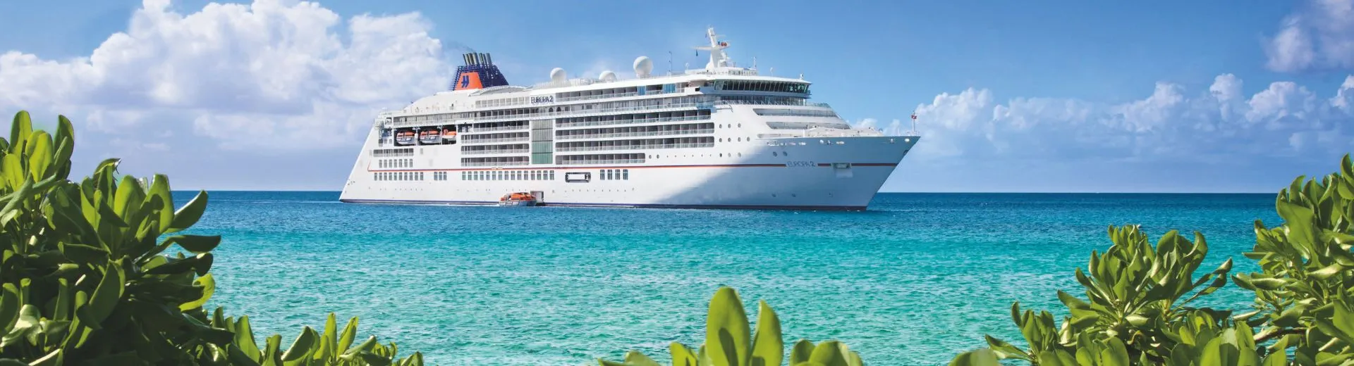 Het luxe cruiseschip MS Europa 2 van Hapag-Lloyd ligt stil voor de kust van een tropisch eiland, met helder turquoise water en weelderig groen op de voorgrond onder een stralend blauwe lucht.
