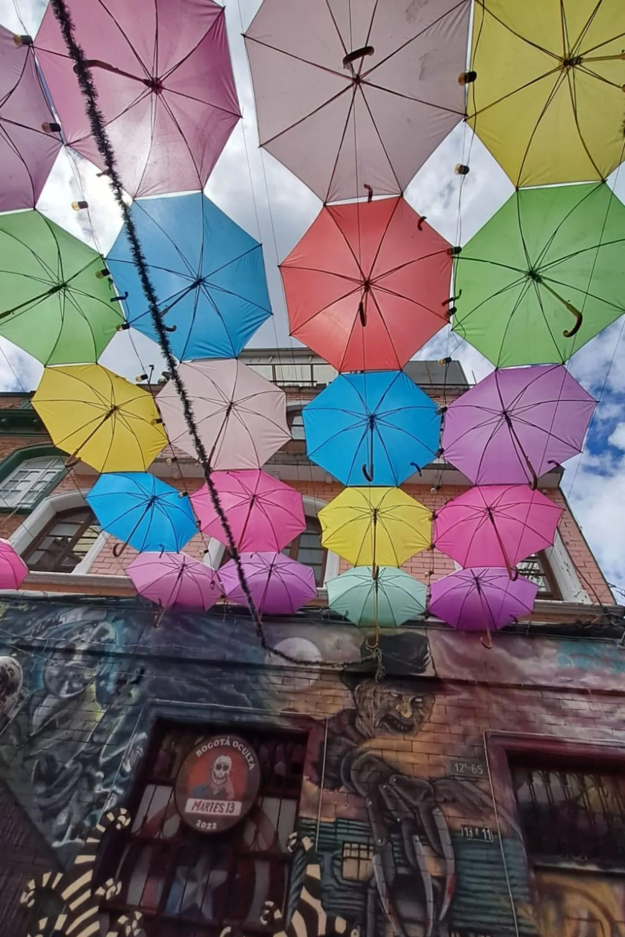Close-up van een plafond van kleurrijke paraplu’s boven een straat, met muurschilderingen en een bord van een café op de achtergrond.