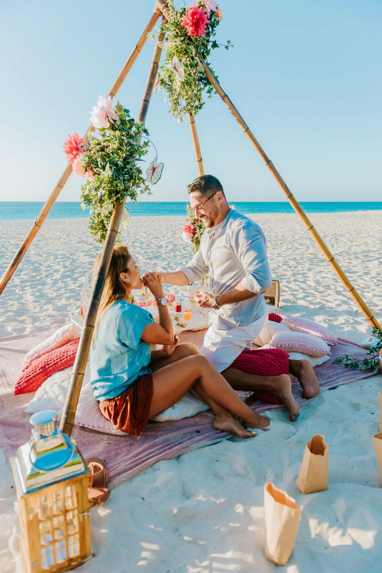 Een close-up van een verliefd stel dat samen geniet van een romantische picknick op een wit zandstrand. Ze zitten dicht bij elkaar, omringd door kaarsen en een prachtig versierde opstelling van bloemen en lekkernijen. De zon schijnt helder, met de azuurblauwe zee en lucht op de achtergrond, wat een warme en intieme sfeer creëert