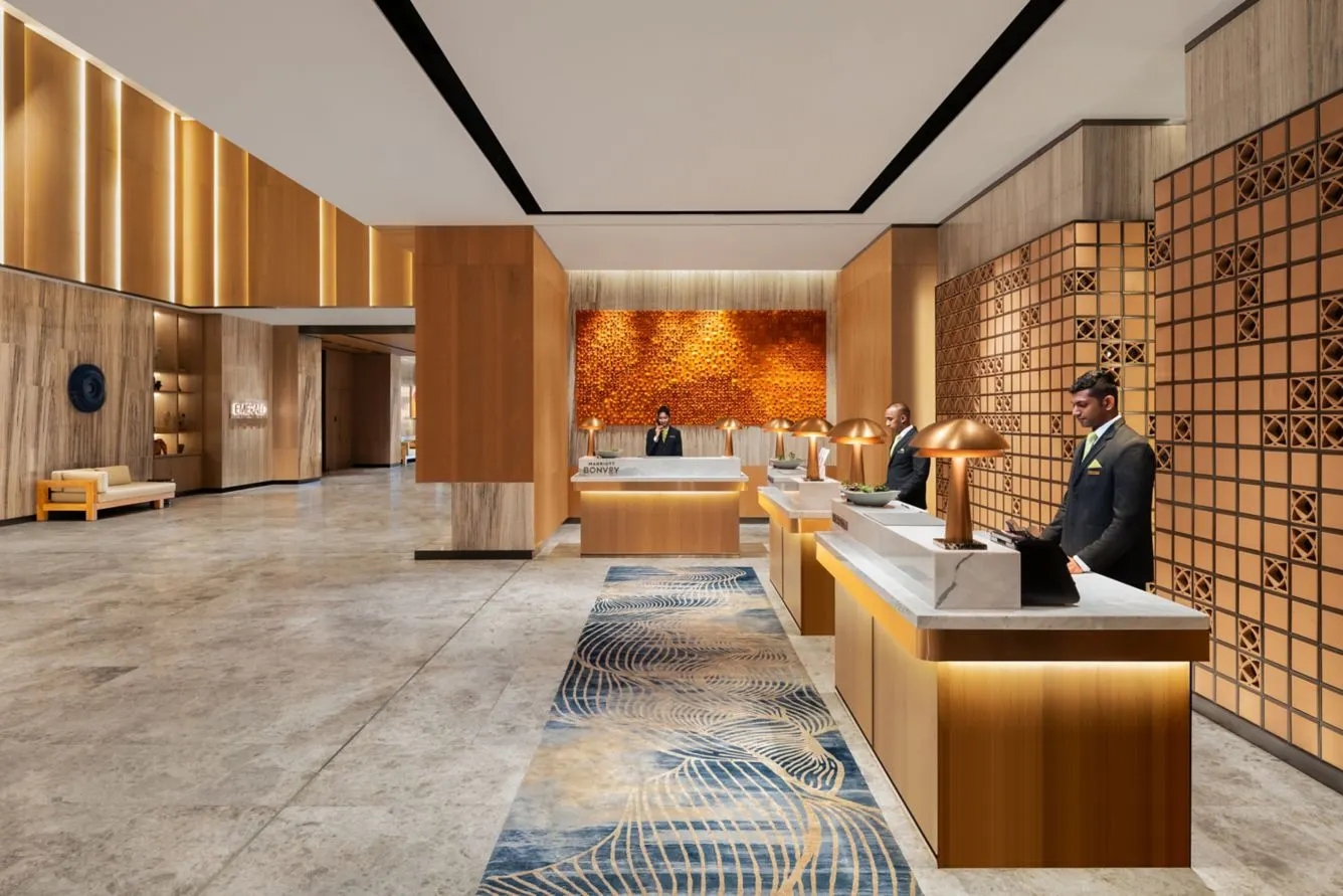 Luxe hotelreceptie van Courtyard by Marriott Colombo met moderne houten accenten, marmeren vloeren en elegante verlichtingsdetails. Drie baliemedewerkers verwelkomen gasten in een ruime, stijlvolle lobby.