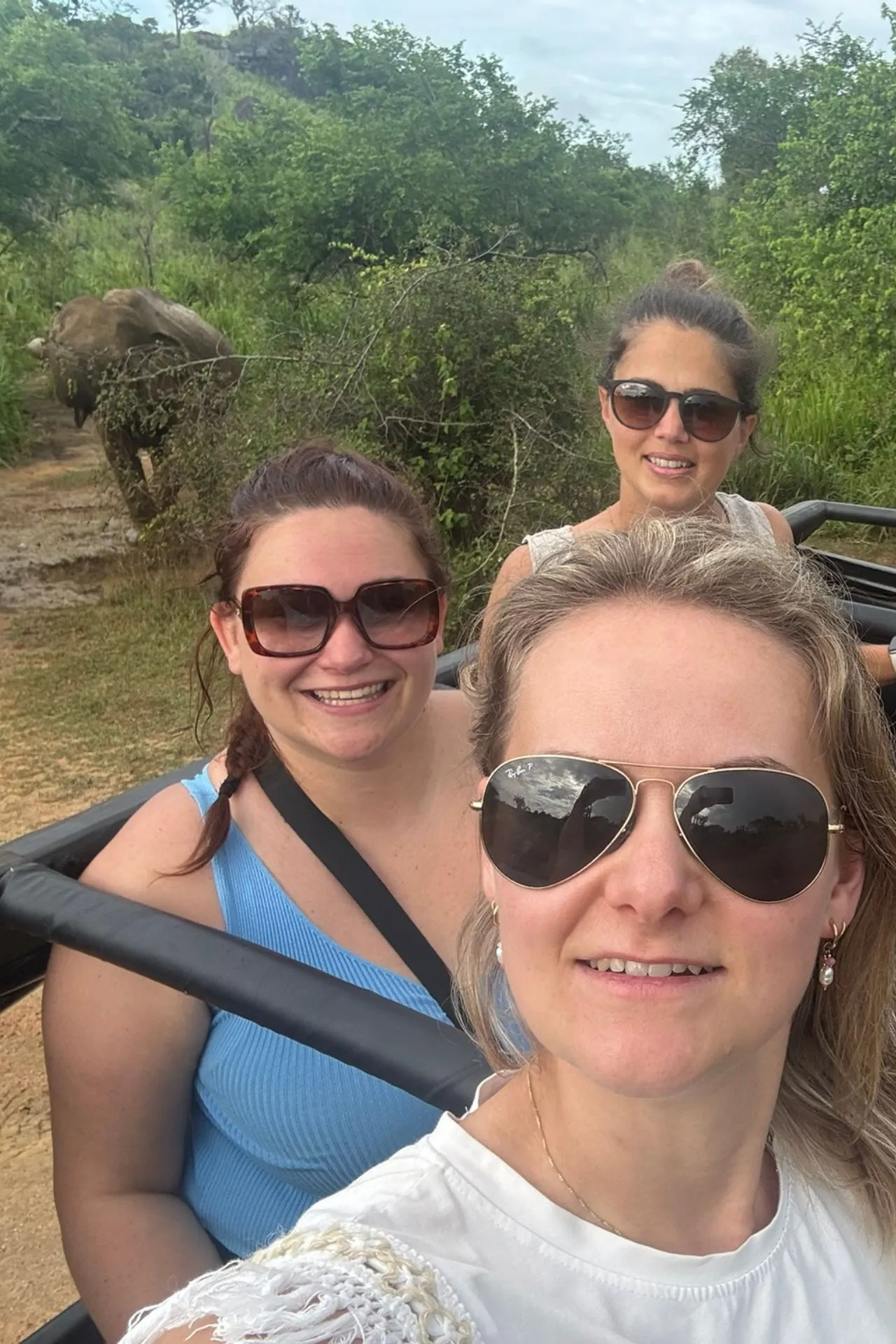 Drie vrouwen glimlachen terwijl ze in een open jeepsafari door een nationaal park in Sri Lanka rijden. Op de achtergrond wandelt een olifant tussen het groen, wat het moment een avontuurlijke en opwindende sfeer geeft. De dames dragen zonnebrillen en zomerse kleding, klaar voor een dag vol natuurschoon en wilde dieren.