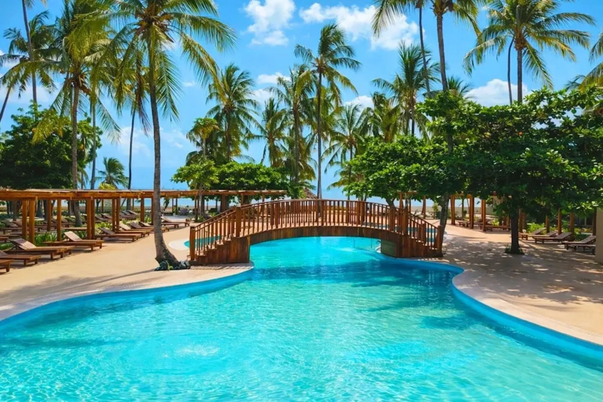 Zwembad van Jaz Amaluna Beach Resort op Zanzibar, omringd door tropische palmbomen en zonneterrassen. Dit stijlvolle adults only resort in Uroa Beach combineert rust, luxe en een kleinschalige sfeer. De groene tuin, houten brug en ontspannen omgeving maken het een ideale plek voor een strandvakantie op Zanzibar.