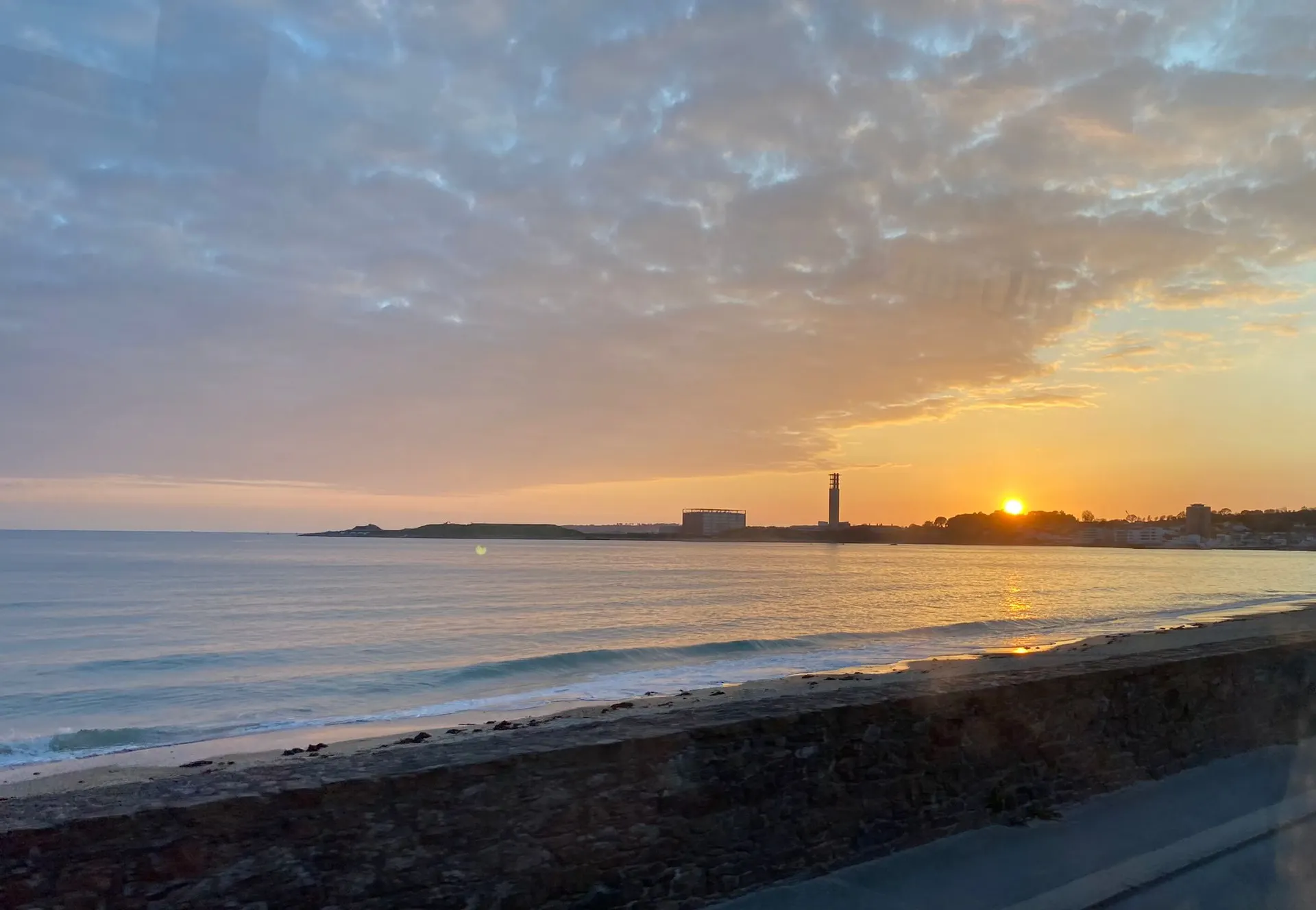 Adembenemende zonsondergang boven de oceaan met een warme oranje gloed die weerspiegelt op het kalme water. Aan de horizon staat een slanke vuurtoren naast een kustlijn met gebouwen, terwijl de lucht gevuld is met zachte wolken die het licht prachtig verspreiden. Perfect voor fotografie, romantische wandelingen langs de kust en vakanties aan zee, met een serene sfeer en panoramisch uitzicht op de ondergaande zon.