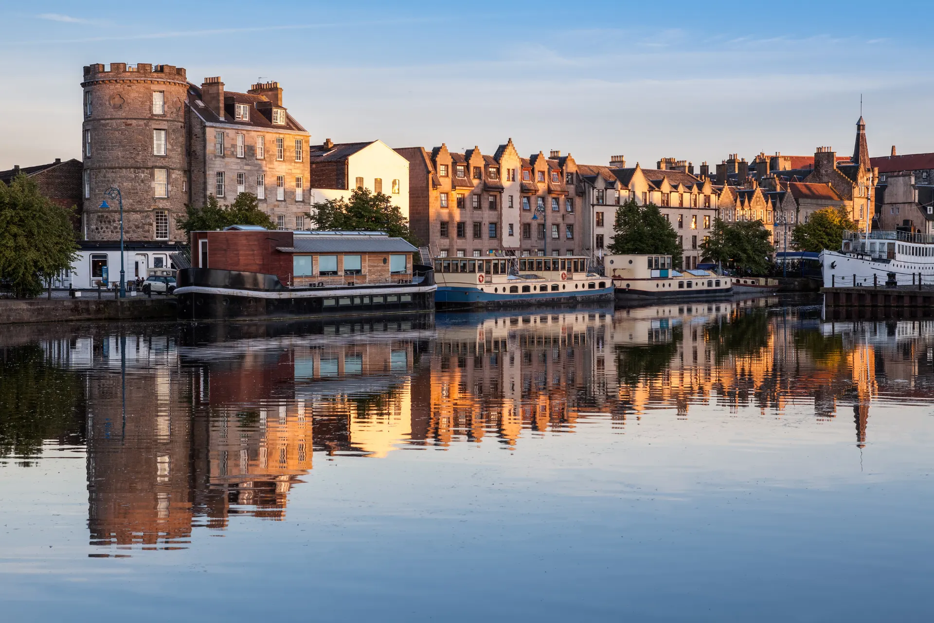 "Historische waterkant met stenen gebouwen en torens weerspiegeld in een rustige rivier, omringd door groene bomen en heldere blauwe lucht. Charmant stadsbeeld ideaal voor stedentrips, cultuurreizen en fotografie. Ontdek pittoreske architectuur, erfgoed en unieke reiservaringen in Schotland. Perfecte bestemming voor liefhebbers van geschiedenis, natuur en romantische uitzichten langs het water."
