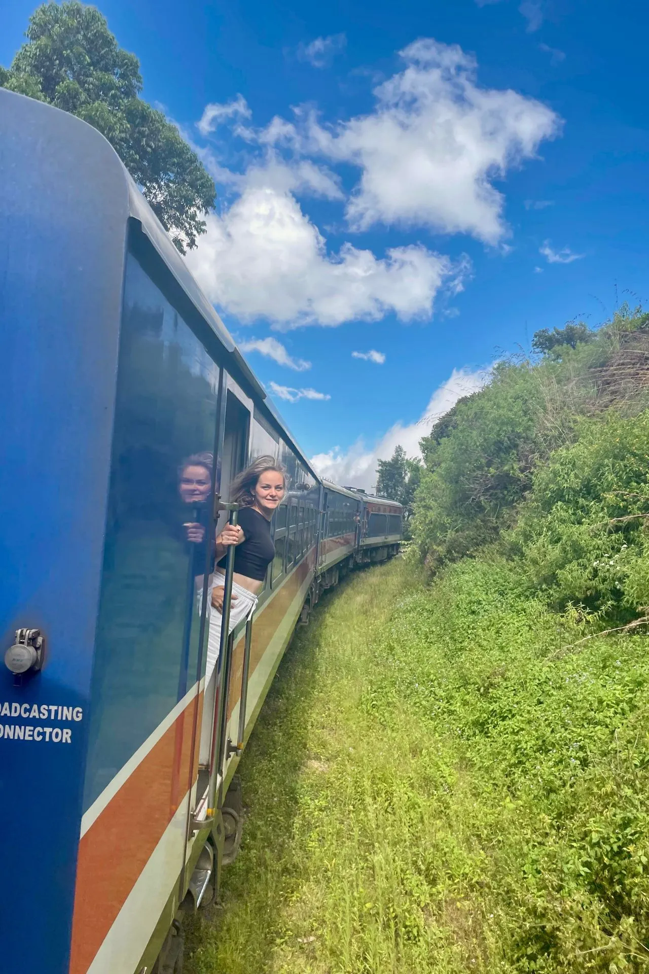 Een vrouw leunt lachend uit het raam van een rijdende trein terwijl deze door het glooiende, groene landschap van Sri Lanka slingert. De blauwe lucht met witte wolken en het contrast van de trein tegen het frisse groen zorgen voor een gevoel van ultieme vrijheid en reisplezier. Dit iconische treinmoment vangt de magie van de wereldberoemde route van Kandy naar Ella.