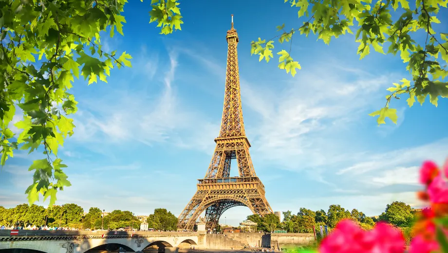 Iconisch uitzicht op de Eiffeltoren in Parijs, omringd door groene bomen en kleurrijke bloemen onder een stralend blauwe lucht. De beroemde toren staat majestueus boven de stad en weerspiegelt de charme en romantiek van Frankrijk. Op de voorgrond zorgen frisse bladeren en levendige tinten voor een zomerse sfeer, terwijl de brug over de Seine elegant in beeld komt. Perfect voor een stedentrip naar Parijs, vol cultuur, architectuur en onvergetelijke bezienswaardigheden in het hart van Frankrijk.