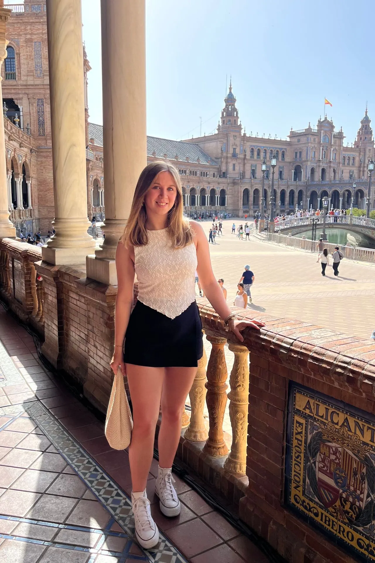 Jonge vrouw poseert lachend op het balkon van Plaza de España in Sevilla, Spanje. Op de achtergrond zijn de indrukwekkende gebouwen van het plein en toeristen op het plein zichtbaar.