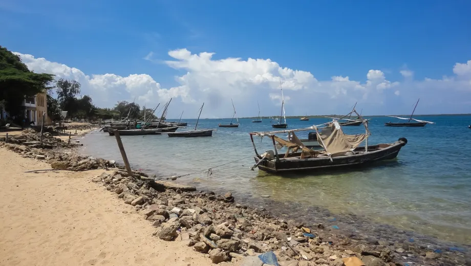 Rustige kust van Kenia met een zandstrand, helderblauw water en kleurrijke bootjes op de horizon.