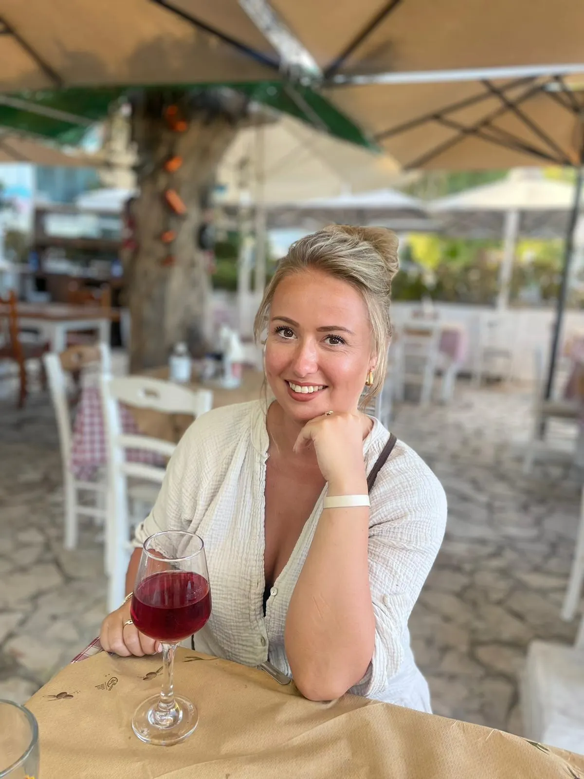 Blonde vrouw glimlacht op een terras met een glas rode wijn in haar hand.