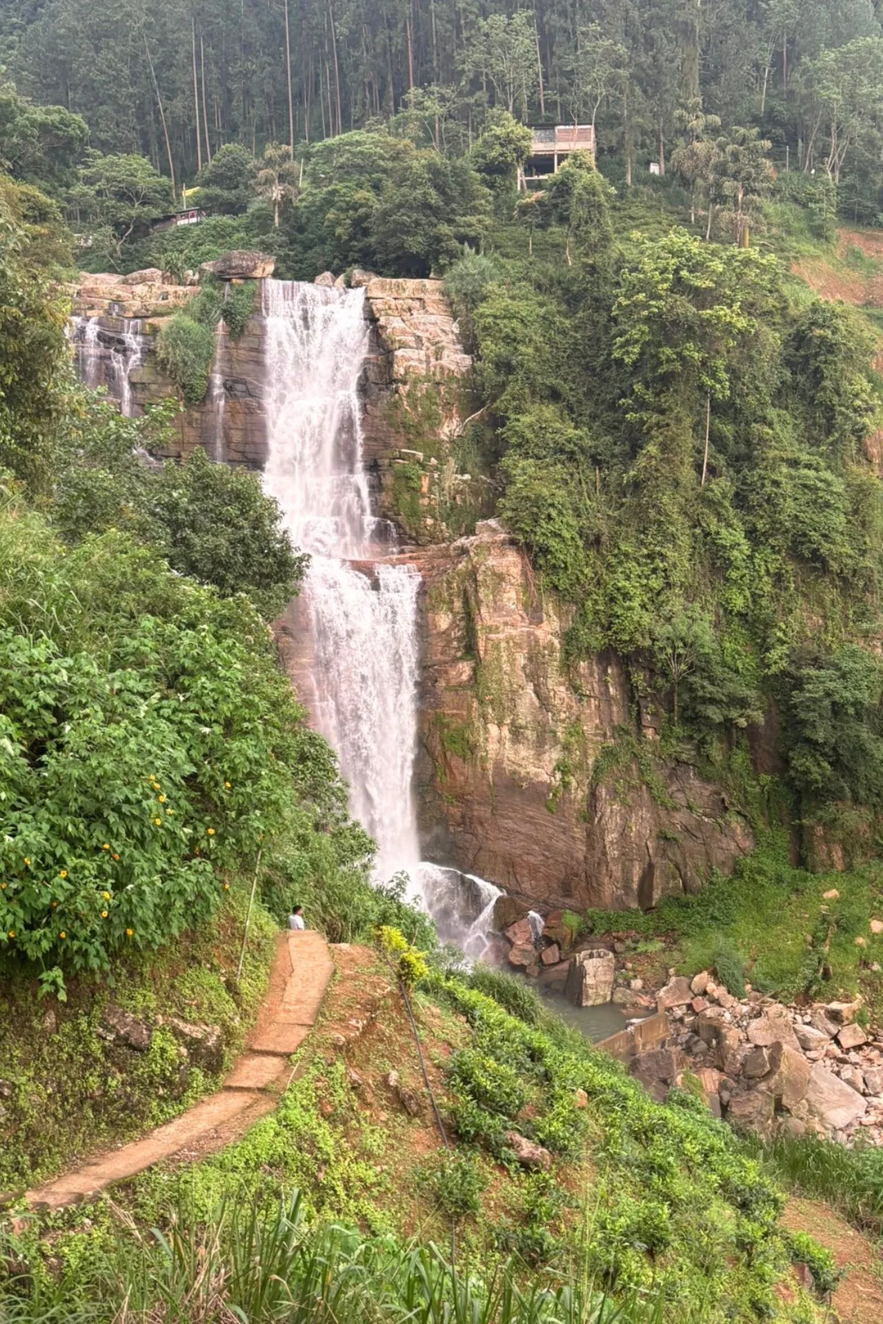 Een hoge waterval stort naar beneden langs een steile rotswand, omringd door dichte, tropische begroeiing. Aan de voet van de waterval stroomt het water tussen rotsblokken door, terwijl een pad links langs de waterval omhoog slingert.