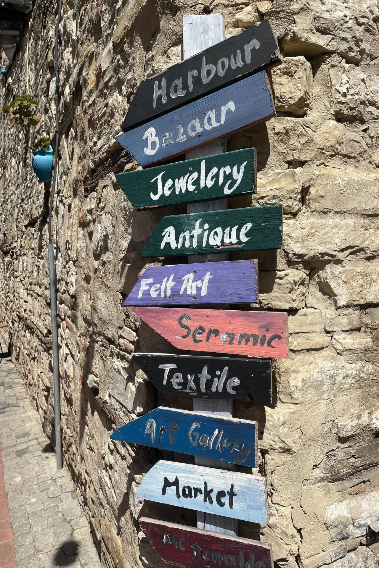 Houten wegwijzer tegen een stenen muur in Kuşadası, met handgeschilderde richtingen naar bazaar, haven en lokale winkels.
