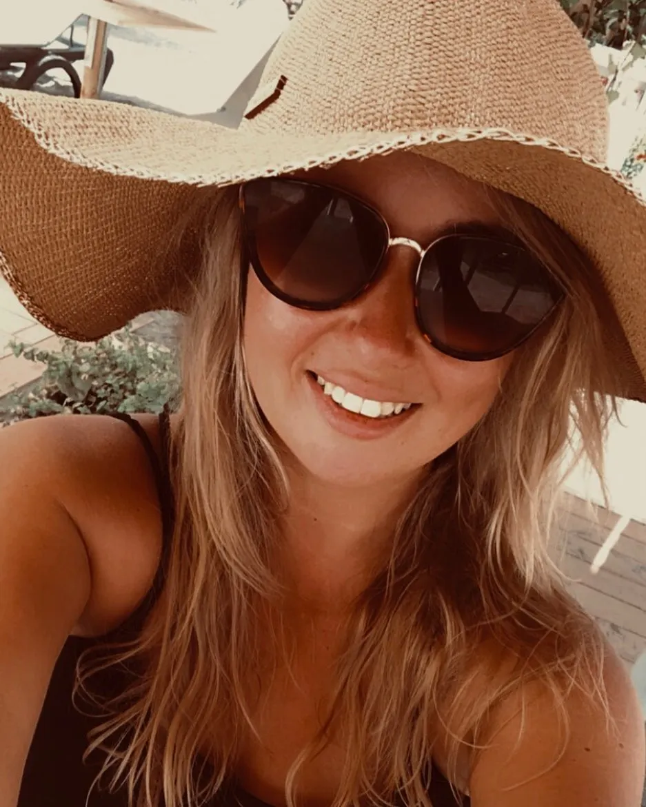 Lachende blonde vrouw met een bruine ronde zonnebril en een strandhoed maakt een selfie.