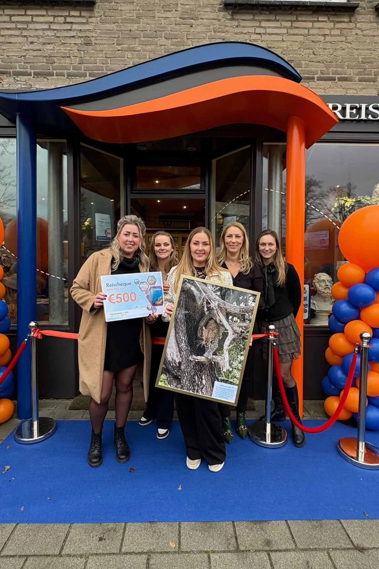 Groep mensen bij de entree van Travelcenter de Wit Asten, omringd door oranje en blauwe ballonnen. Eén persoon houdt een grote foto van een luipaard vast, naast een cheque van €500,- voor de fotowedstrijd.