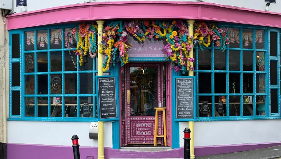 Opvallende kleurrijke gevel van een restaurant of café met felroze en turquoise tinten, grote ramen en een deur versierd met bloemen en slingers in levendige kleuren. Aan weerszijden van de ingang staan zwarte borden met menu-informatie, wat een uitnodigende en creatieve uitstraling geeft. Deze unieke locatie is ideaal voor liefhebbers van bijzondere eetervaringen, Instagram-waardige hotspots en sfeervolle restaurants in een levendige straatomgeving.