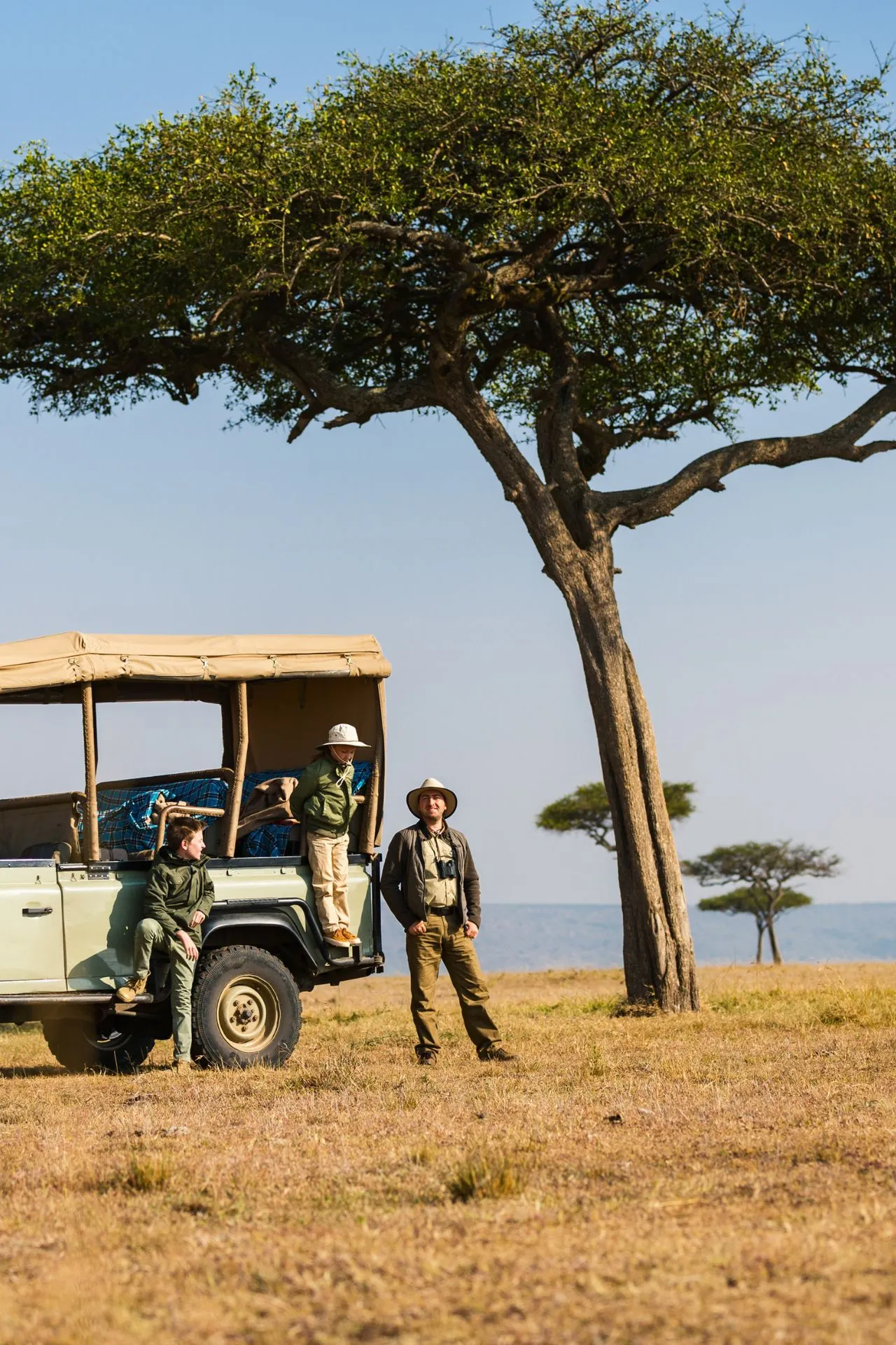 Een uitgestrekt woestijnlandschap in de Sahara, Afrika, met hier en daar een enkele boom en een verder vlak uitzicht. Op de voorgrond staat een jeep met een man die turend de omgeving verkent, terwijl twee kinderen uit de jeep klimmen om de woestijn te ontdekken.