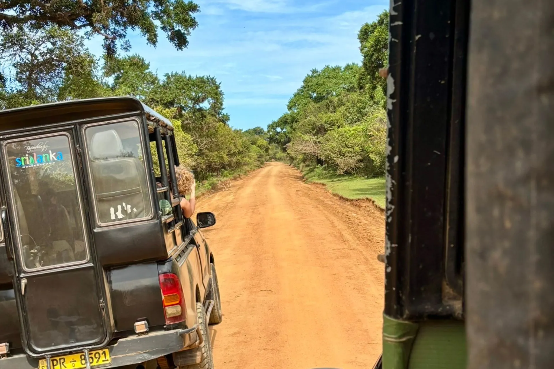 Een jeep rijdt over een oranje zandweg door Yala National Park, Sri Lanka. Een passagier met krullend blond haar leunt uit het raam om foto's te maken. De omgeving bestaat uit weelderig groen en bomen onder een heldere blauwe lucht.