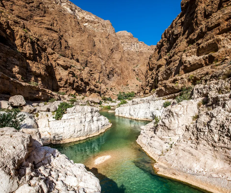 Indrukwekkende kloof in Oman met steile rotswanden in warme aardetinten en een smaragdgroene rivier die zich door het landschap slingert. Het heldere water weerspiegelt de blauwe lucht, terwijl witte kalksteenformaties contrasteren met de ruige bergen. Deze natuurlijke oase is een populaire bestemming voor avontuurlijke reizigers die houden van wandelen, zwemmen en het ontdekken van verborgen parels in het Midden-Oosten, ideaal voor een unieke en authentieke reiservaring.