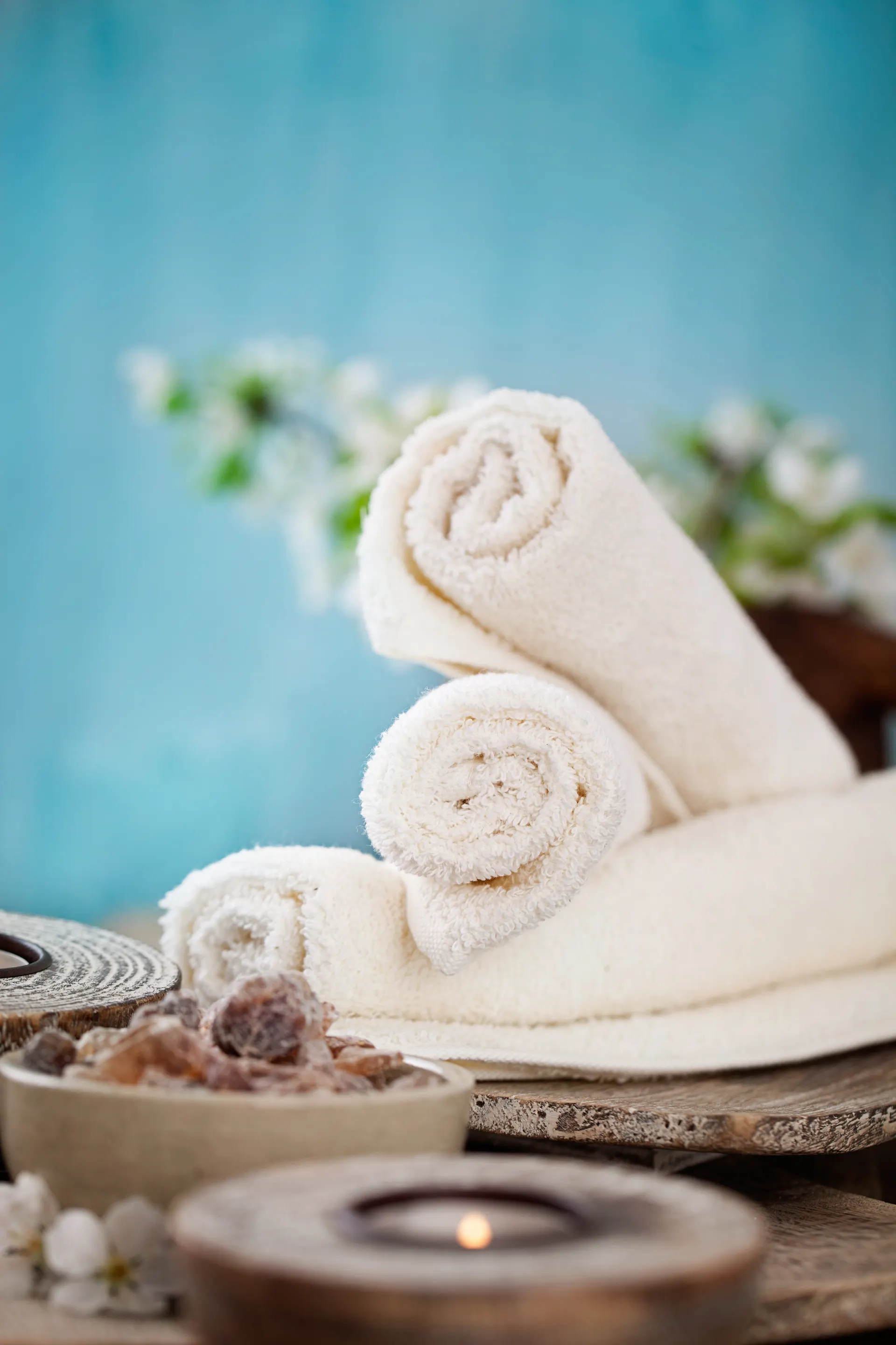 Wellness en ontspanning met opgerolde witte handdoeken op een houten oppervlak, omringd door natuurlijke decoratie zoals stenen, kaarsen en bloemen. Op de achtergrond een zachte blauwe tint die een rustgevende sfeer creëert, ideaal voor een spa-ervaring of beautybehandeling. Perfect voor een luxe wellnessresort of spa in Egypte of een andere zonnige bestemming, gericht op ontspanning, verzorging en premium service.
