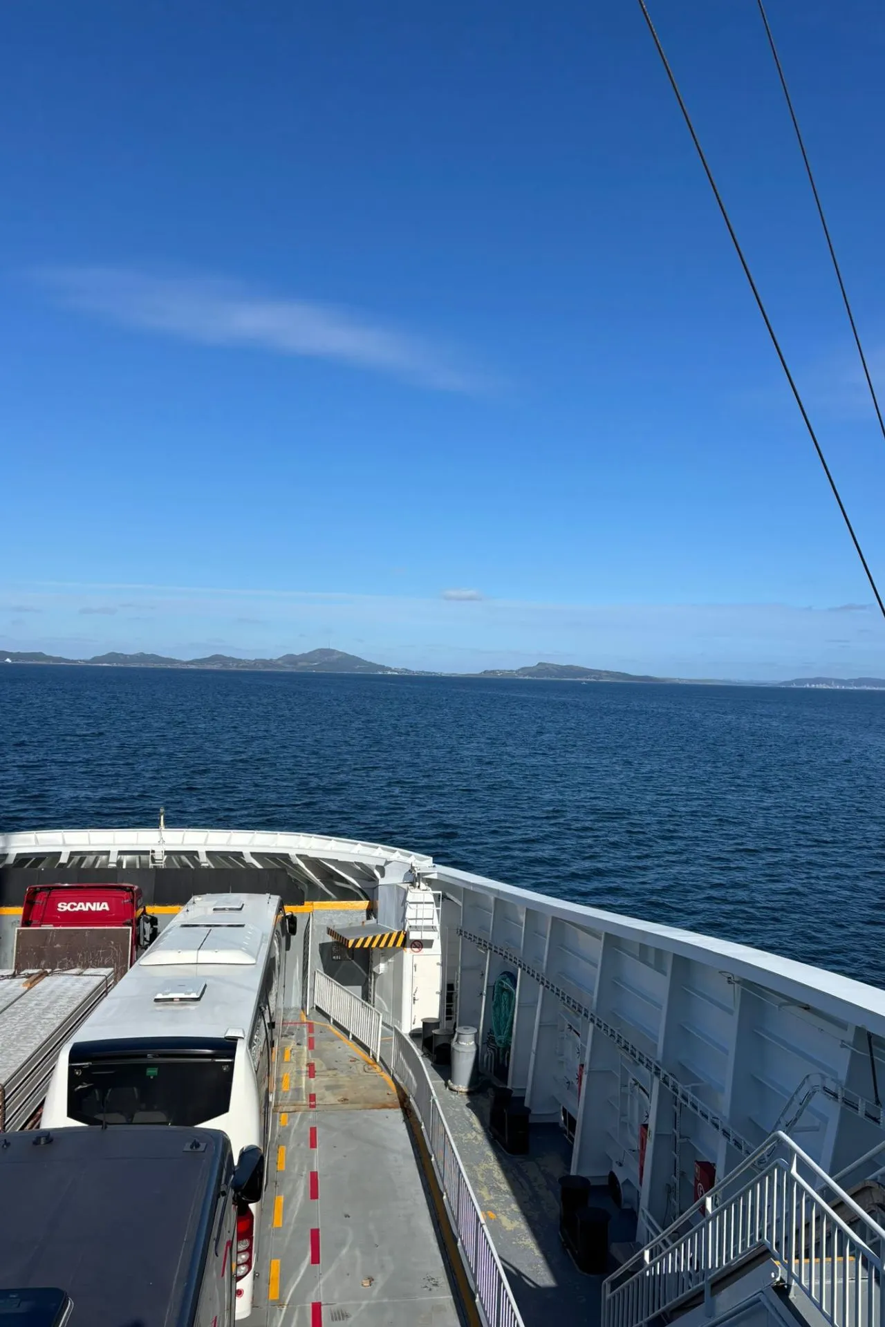 Voorkant van een veerboot met voertuigen op het dek, waaronder een witte bus en een vrachtwagen. De boot vaart op een rustige blauwe zee met lage eilanden in de verte onder een heldere blauwe lucht.