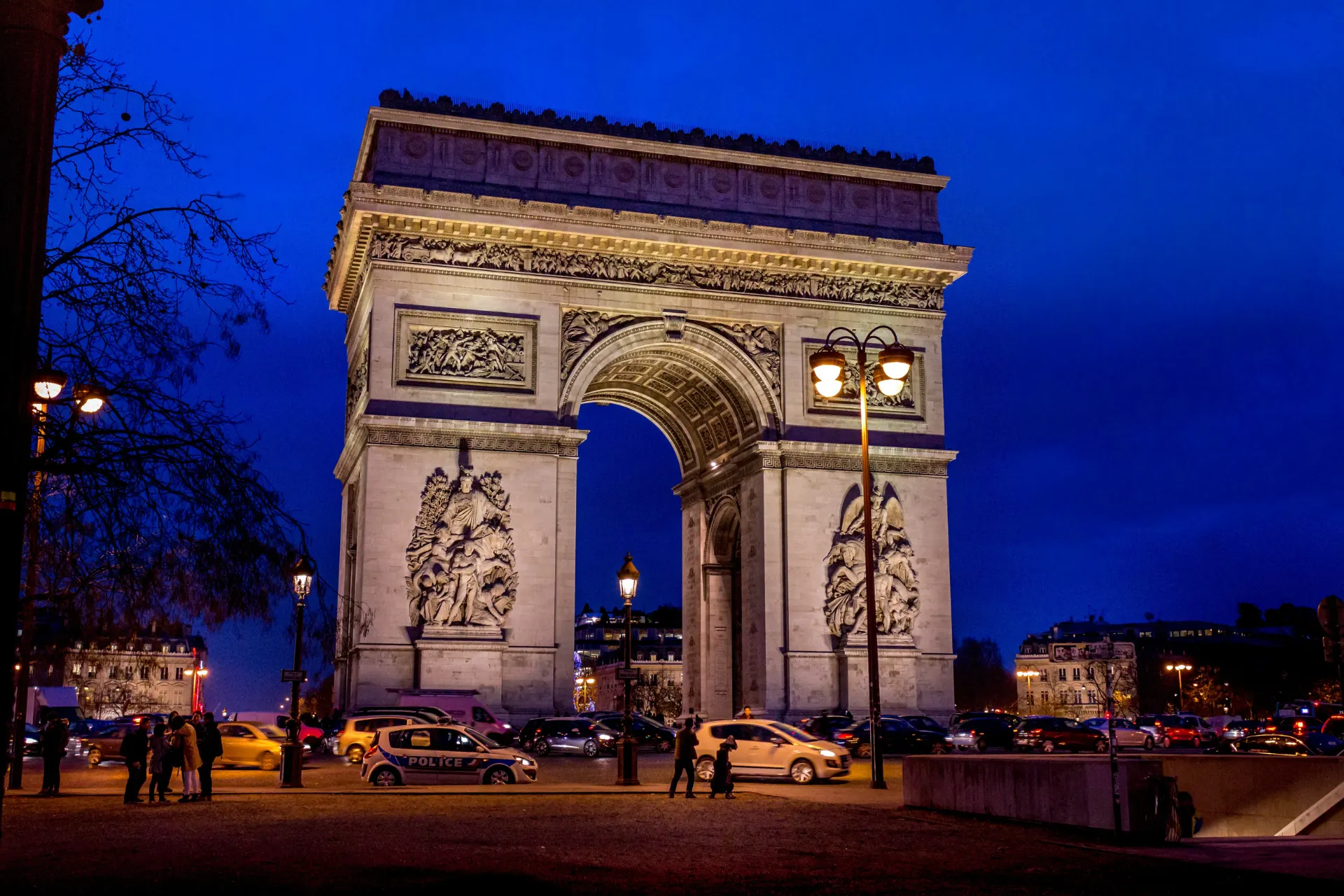 De Arc de Triomphe in Parijs, prachtig verlicht tegen een diepe blauwe avondlucht. Het monument, gelegen aan de beroemde Champs-Élysées, toont gedetailleerde reliëfs en klassieke architectuur. Op de voorgrond rijden auto’s en lopen mensen langs straatlantaarns die warm licht verspreiden. Iconische bezienswaardigheid en historisch symbool van Frankrijk, populair voor toerisme, fotografie en cultuur.

