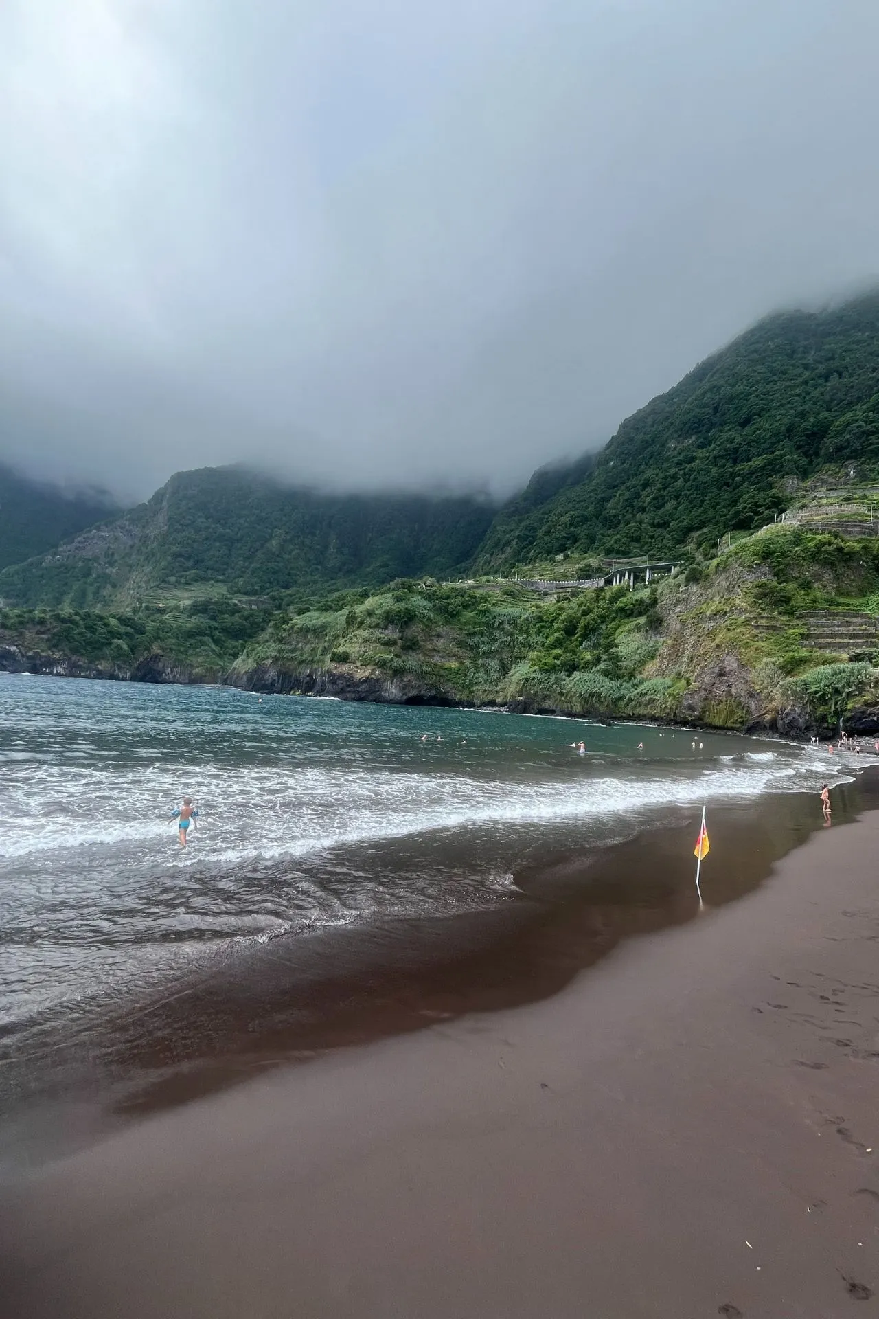 Donkere zandstrand aan de kust van Madeira met golven die op het strand rollen. Op de achtergrond groene bergen deels bedekt door mist en wolken, enkele mensen zwemmen in zee.
