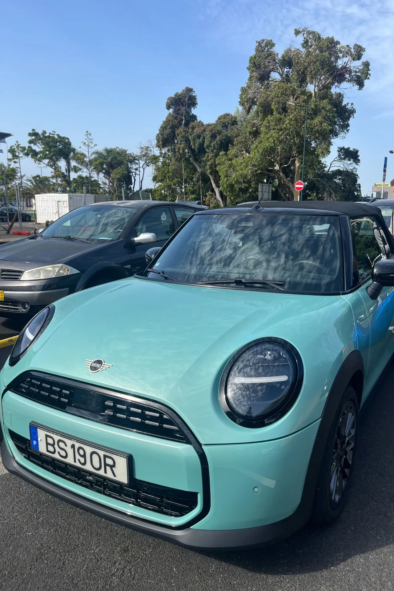 Mintgroene Mini Cooper cabriolet geparkeerd op een asfaltparkeerplaats, met bomen en andere auto’s op de achtergrond. De auto heeft een Europese kentekenplaat