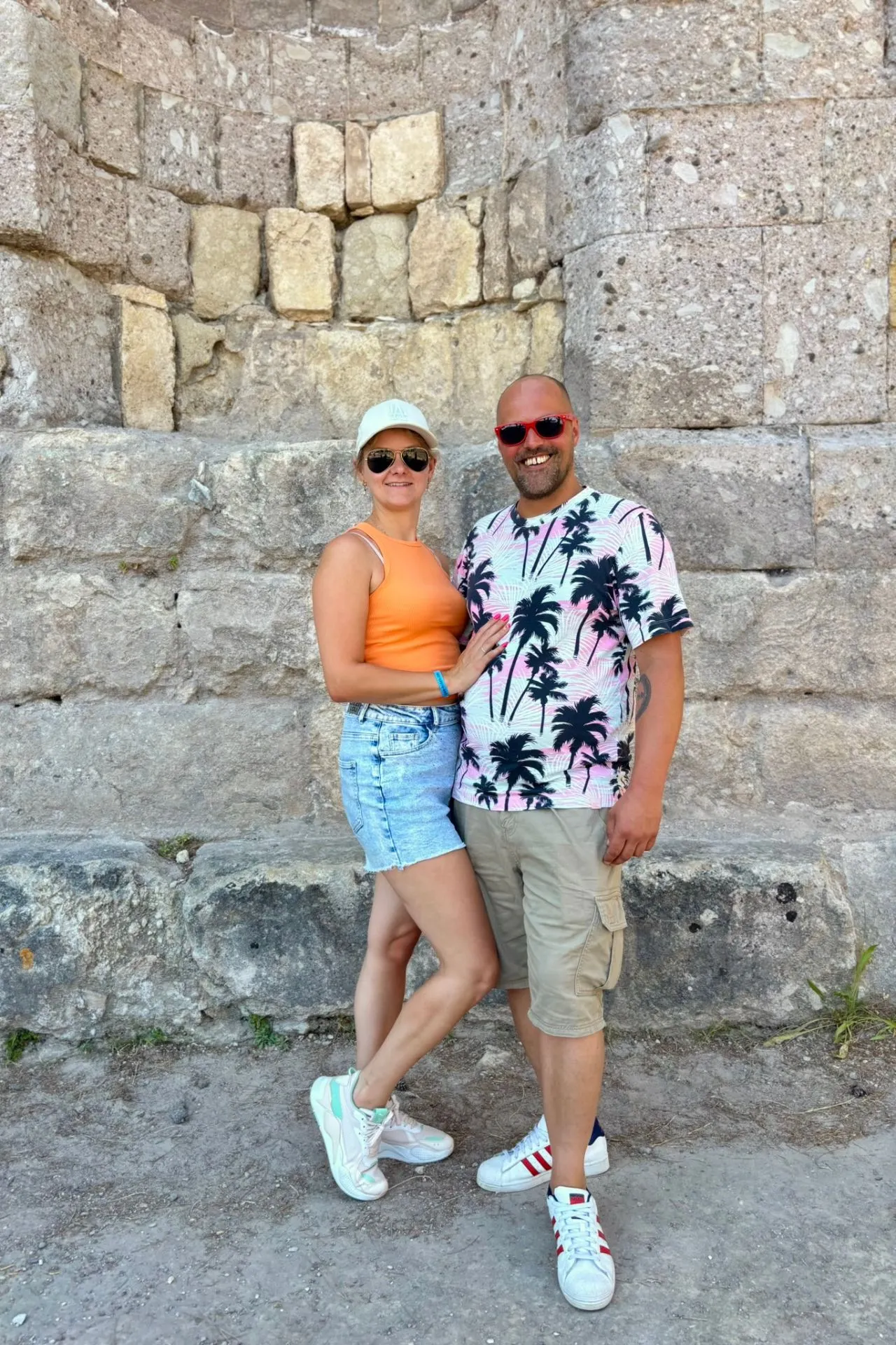 Stel poseert lachend voor een historische stenen muur van het Asklepion op Kos, tijdens een zonnige excursie