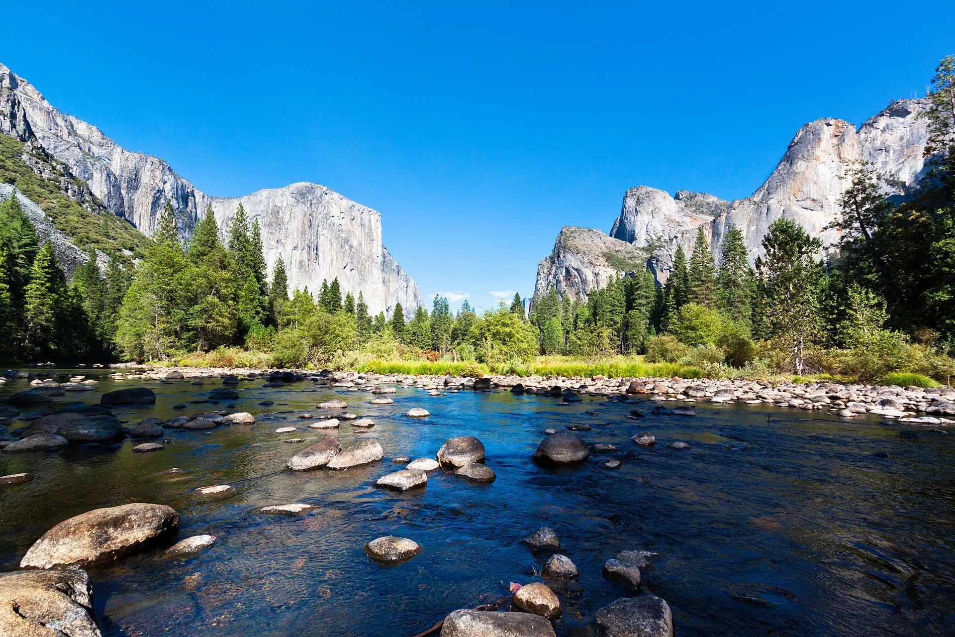 Heldere rivier met rotsblokken en uitzicht op granieten kliffen in Yosemite National Park, Californië. Een iconische bestemming voor natuurliefhebbers en een van de meest bezochte nationale parken in Amerika. Yosemite biedt spectaculaire wandelroutes, watervallen en uitzichten die je Amerika-reis onvergetelijk maken.