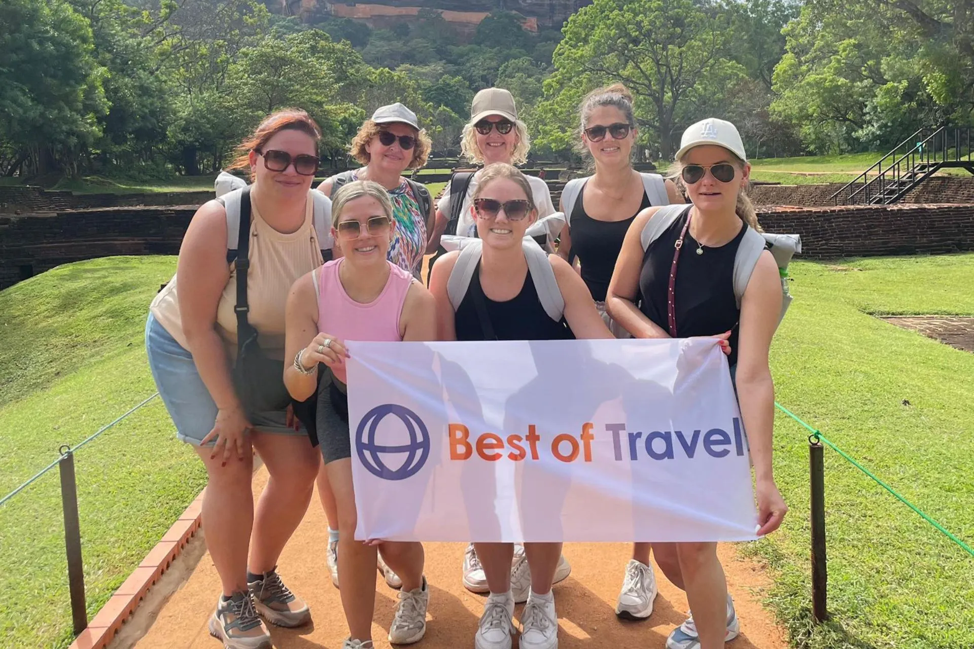 Groepsfoto van zeven lachende reisprofessionals tijdens een studiereis in Sri Lanka, georganiseerd door Best of Travel. De deelnemers poseren op een zonnige locatie met een groen landschap en historische ruïnes op de achtergrond. Ze houden samen een banner vast met het logo van Best of Travel. De sfeer is ontspannen en enthousiast, passend bij een leerzame en inspirerende reiservaring.